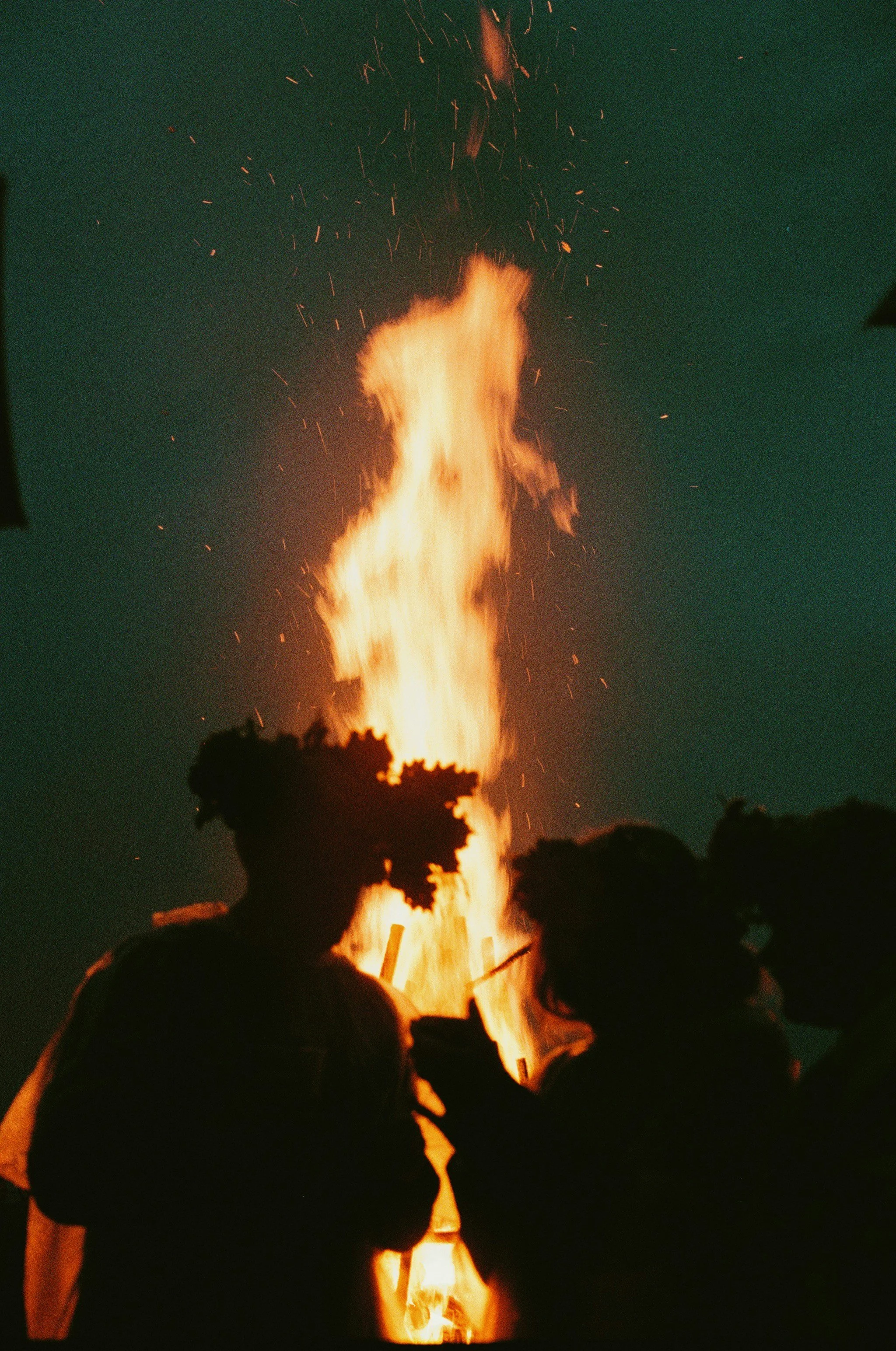 Silhouettes of three people standing near a large bonfire at night.