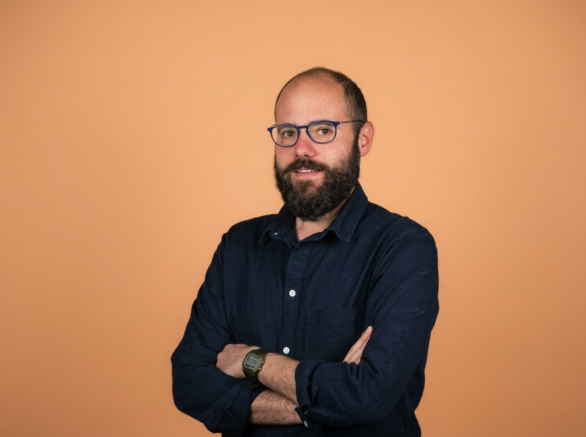 Panagiotis Gkanas - Man with a beard, blue glasses, and a smartwatch, wearing a navy blue shirt, standing against an orange background.