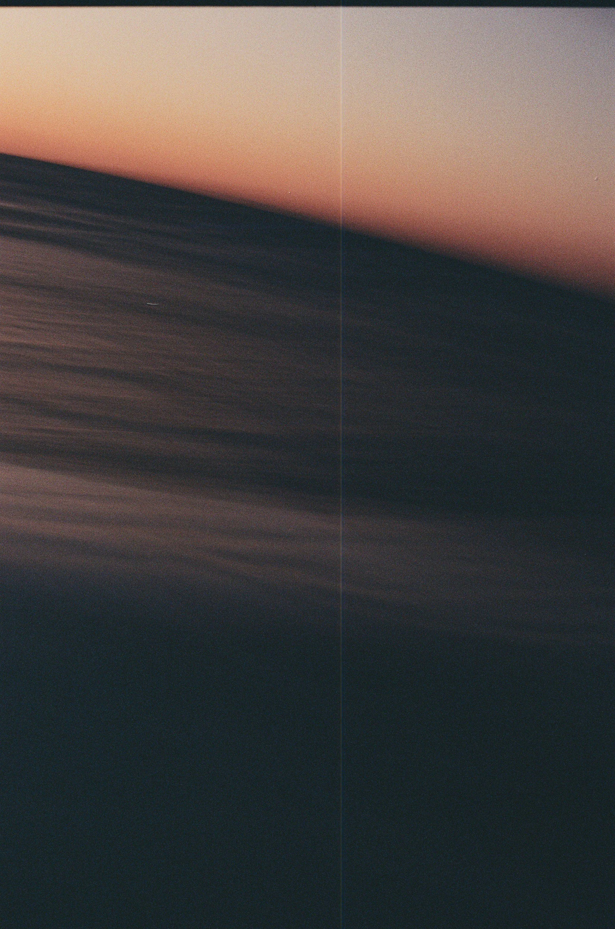 Blurred landscape with a horizon line, showing a sky with gradient colors from light pink to dark purple, and abstract dark shapes in the foreground.