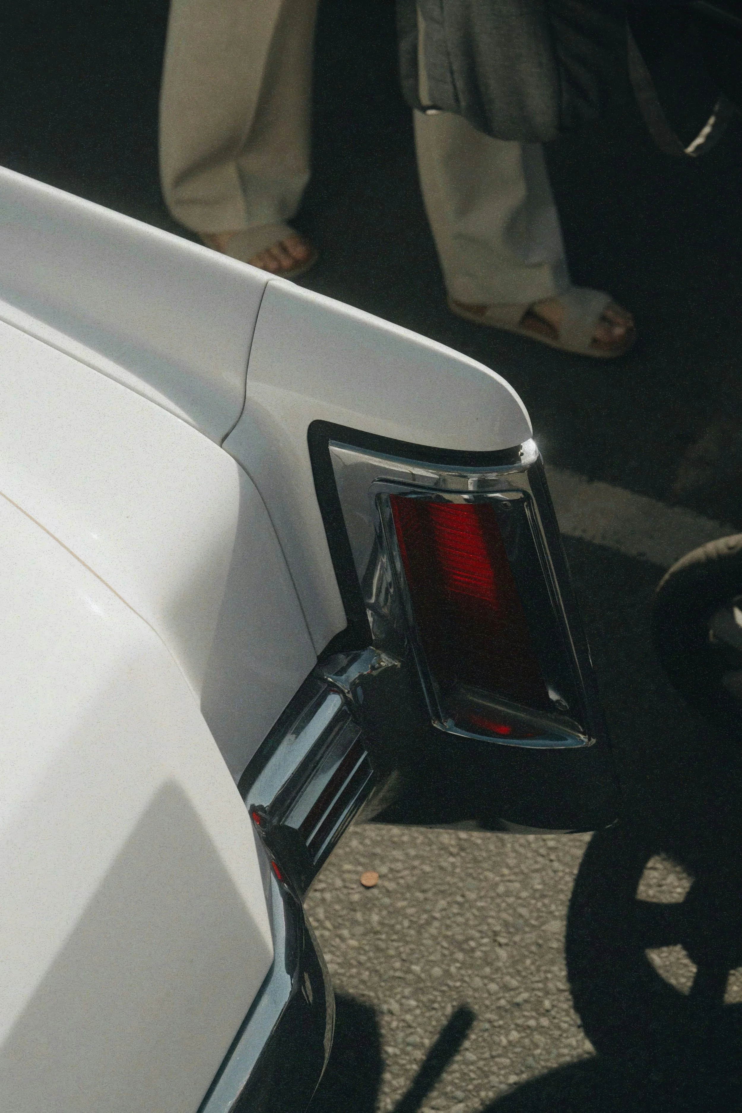 Close-up of the back of a white vintage car showing tail lights and a partial bumper, with people standing nearby.