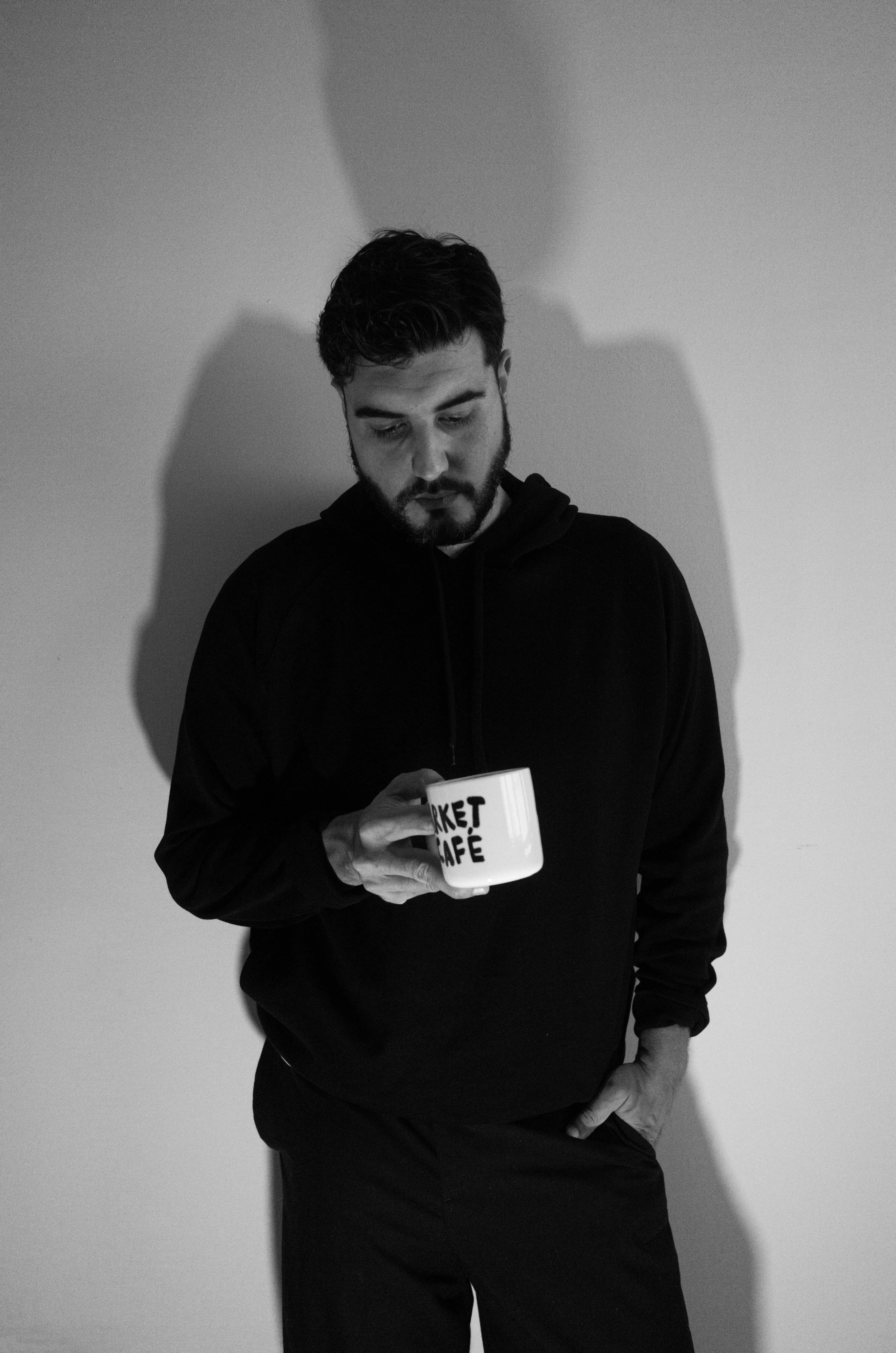 A young man with dark hair and a beard, wearing a black hoodie, stands against a light-colored wall, looking down and holding a coffee cup with the words 'Market Café' printed on it. His shadow is cast on the wall behind him.