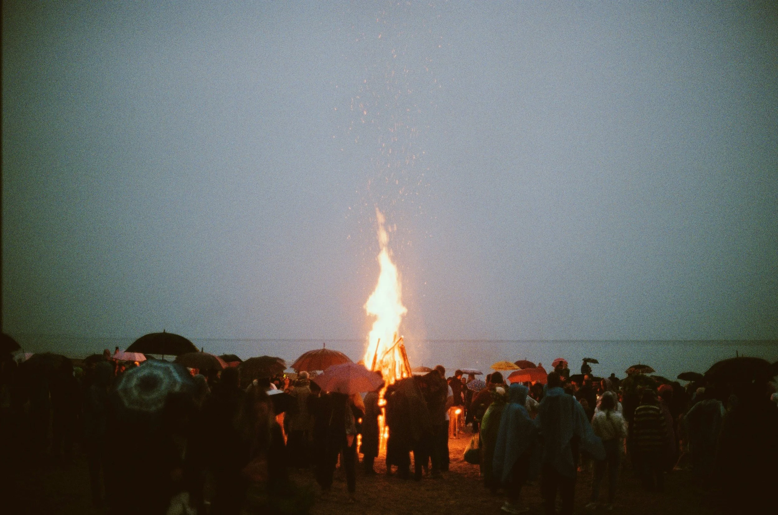 Crowd gathered on a rainy day watching a large bonfire with sparks flying into the sky near the water.