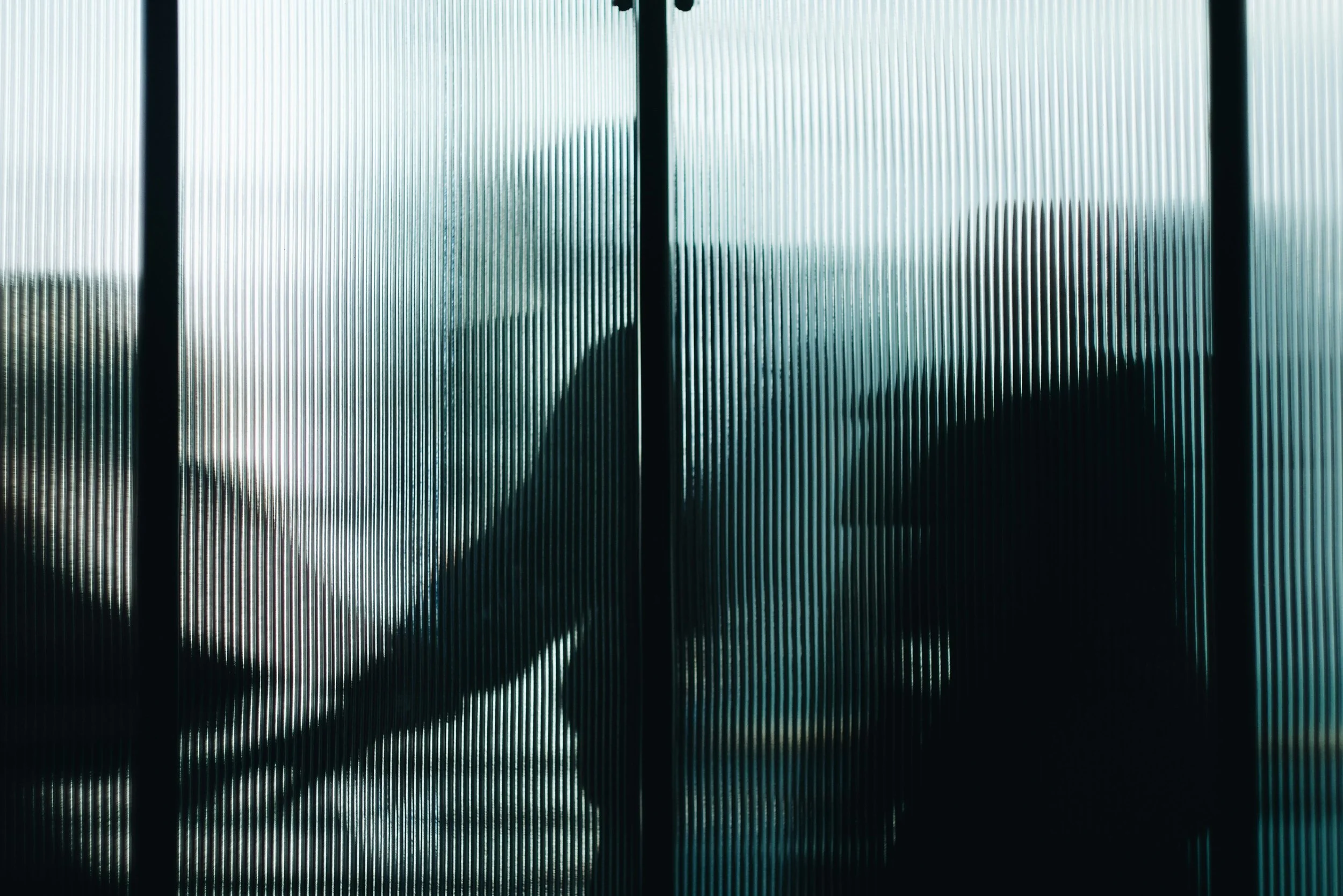 Silhouette of a person with an umbrella seen through ribbed glass panels during rainy weather.