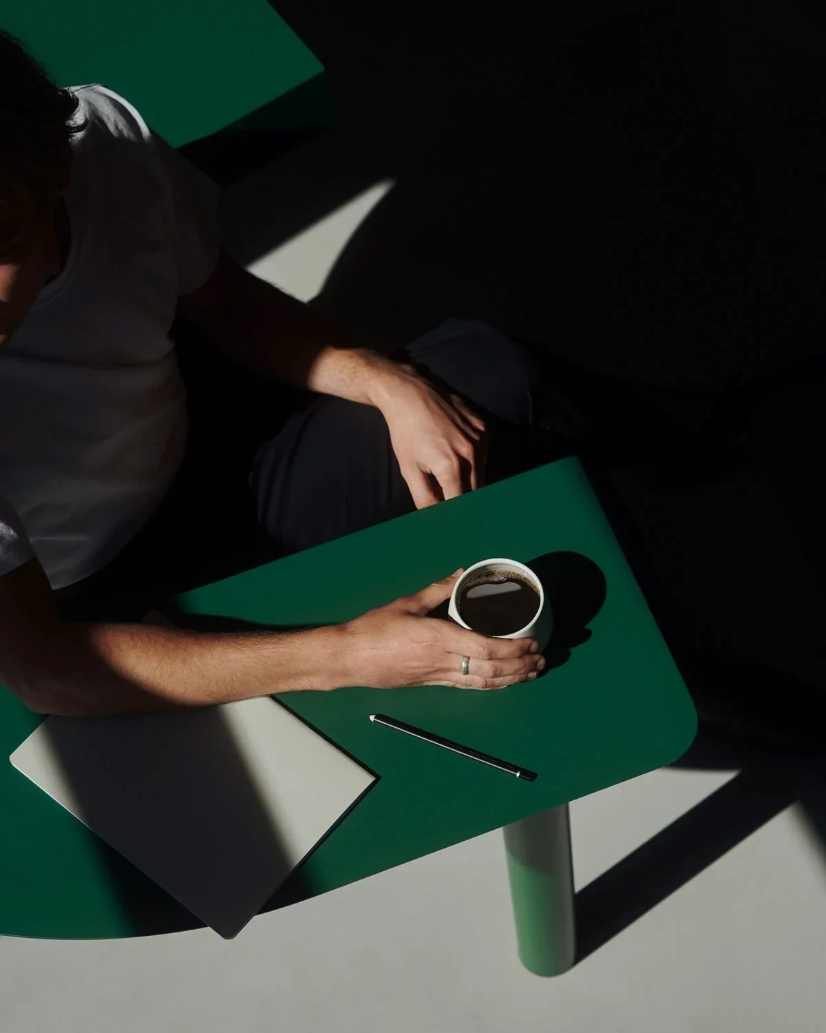 Person holding a cup of black coffee on a green table, with a notepad and pen, and bright sunlight casting shadows.