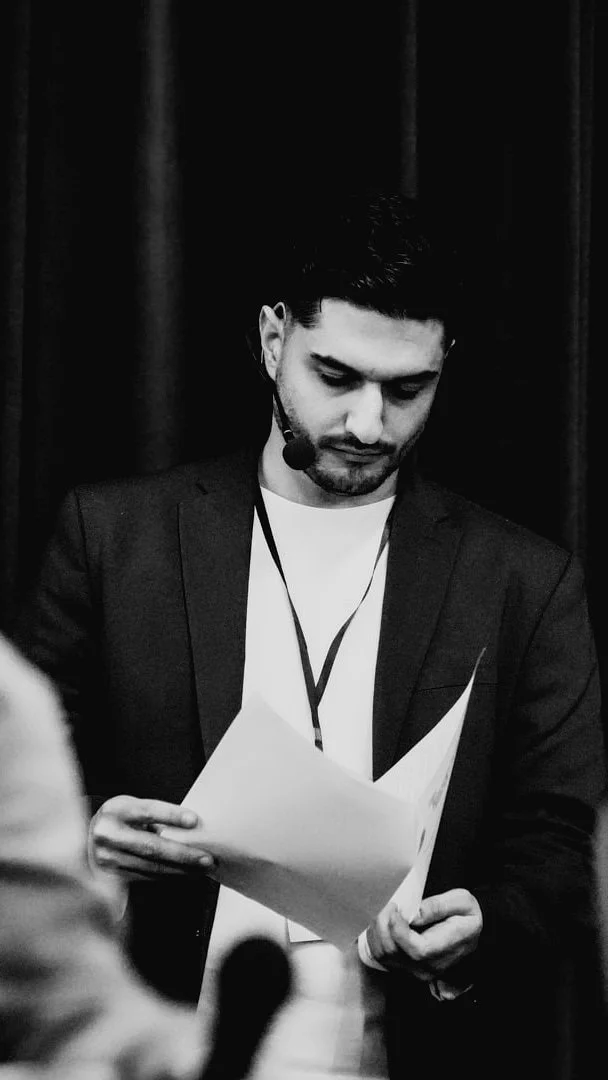 Man with dark hair, beard, and wearing a suit with a headset microphone, looks down at papers in his hands in front of a dark curtain.