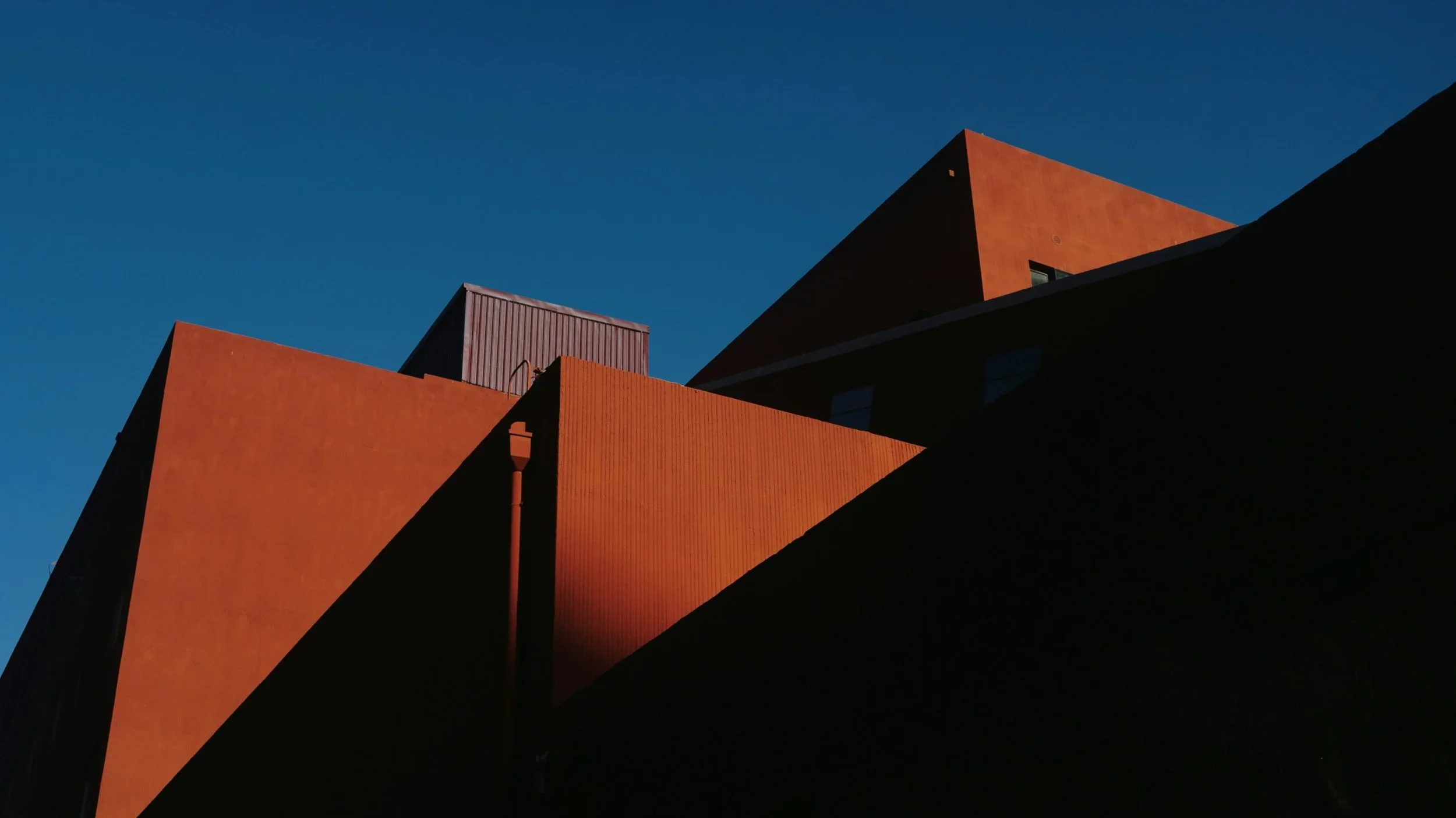 Photo of modern buildings with red and dark-colored walls under a clear blue sky, sunlight casting shadows on the structures.