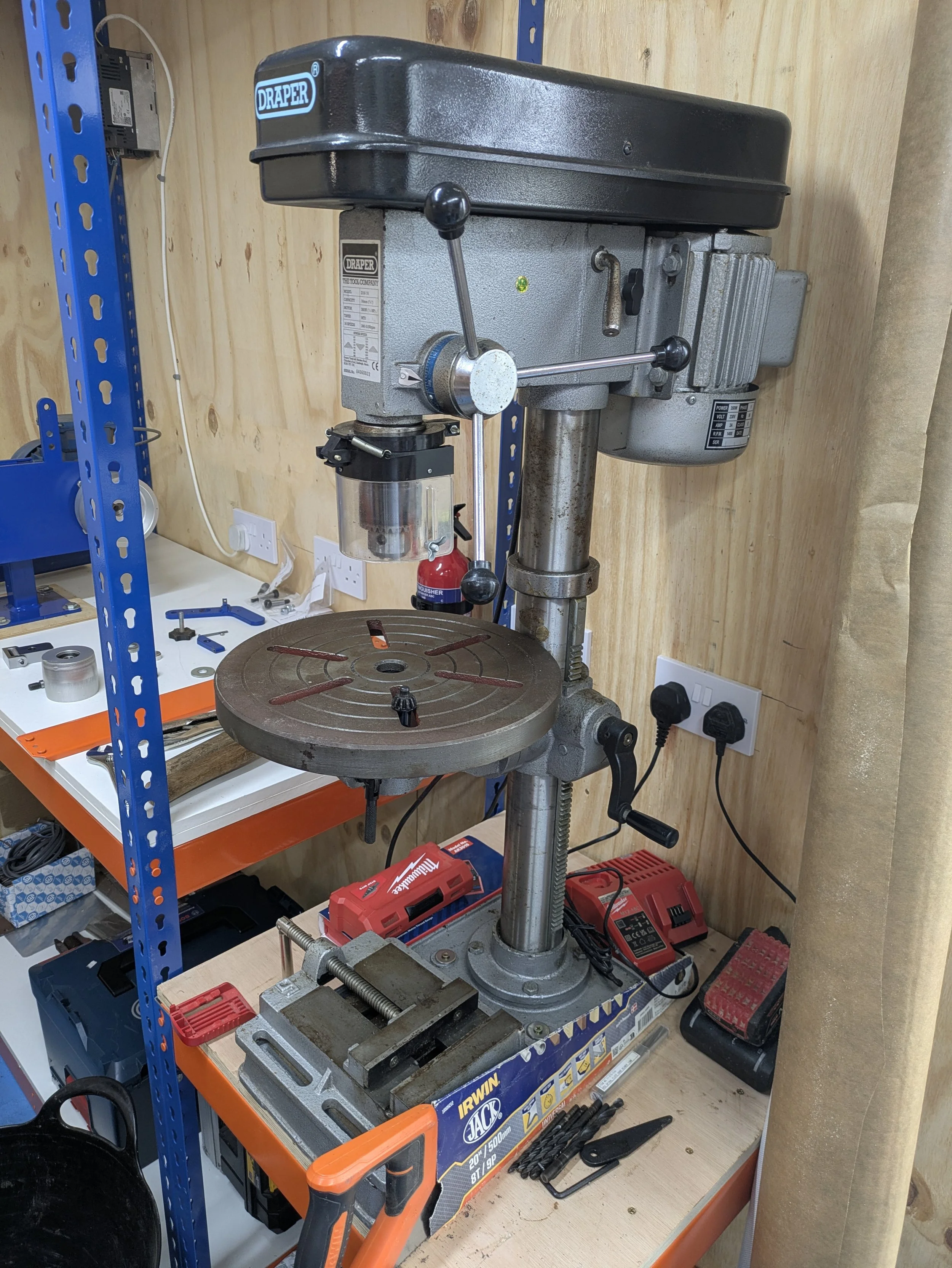 A workshop drill press mounted on a workbench with various tools and equipment surrounding it.