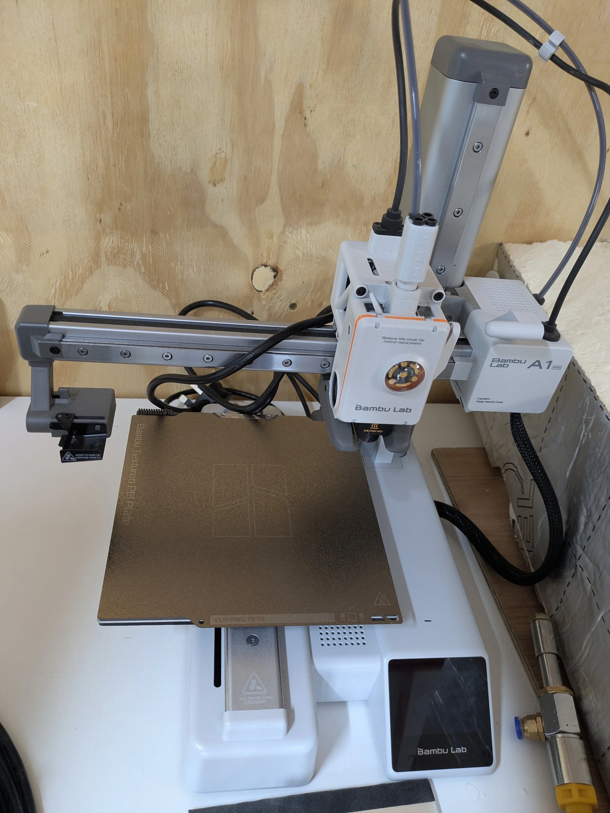 A laboratory setup with a bamboo lab 3D printer on a white surface, a computer monitor, and various cables. The background features a wood-paneled wall with some holes.