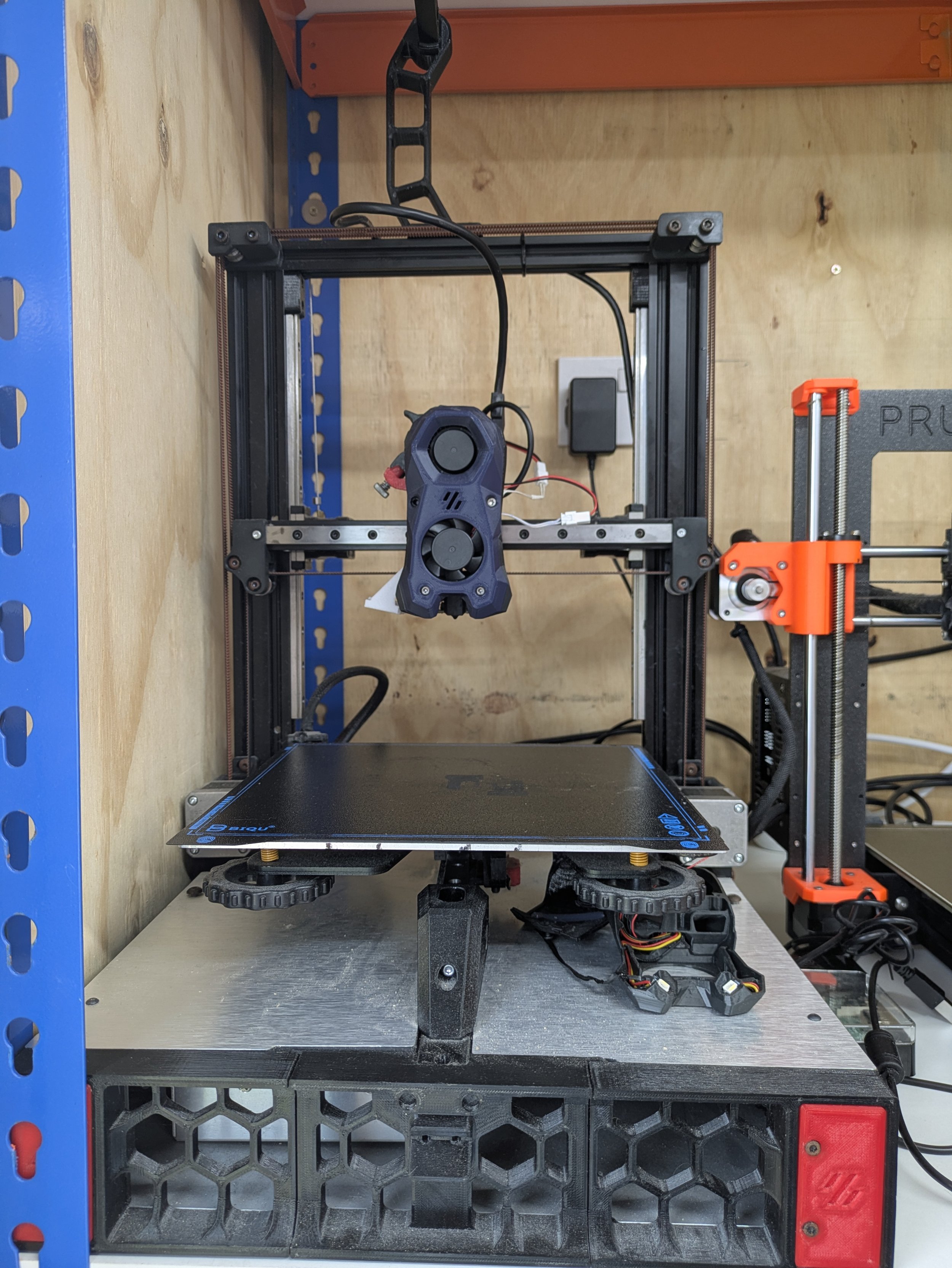 A partially assembled 3D printer on a workbench with various components and tools around it.