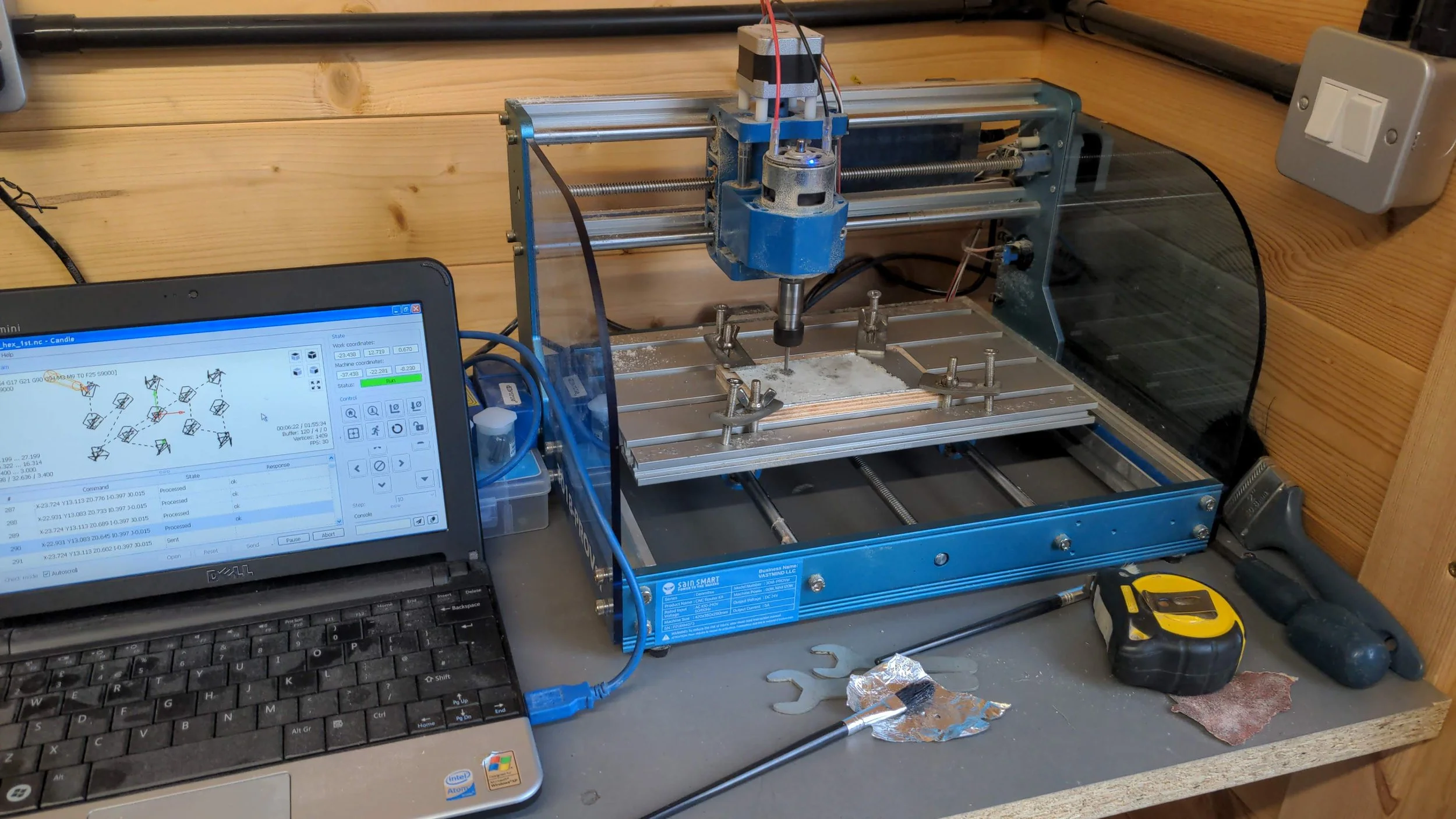 A small CNC machine on a workbench connected to a laptop displaying a design software. The setup includes a yellow tape measure, a pair of pliers, a piece of sandpaper, and some tools nearby.