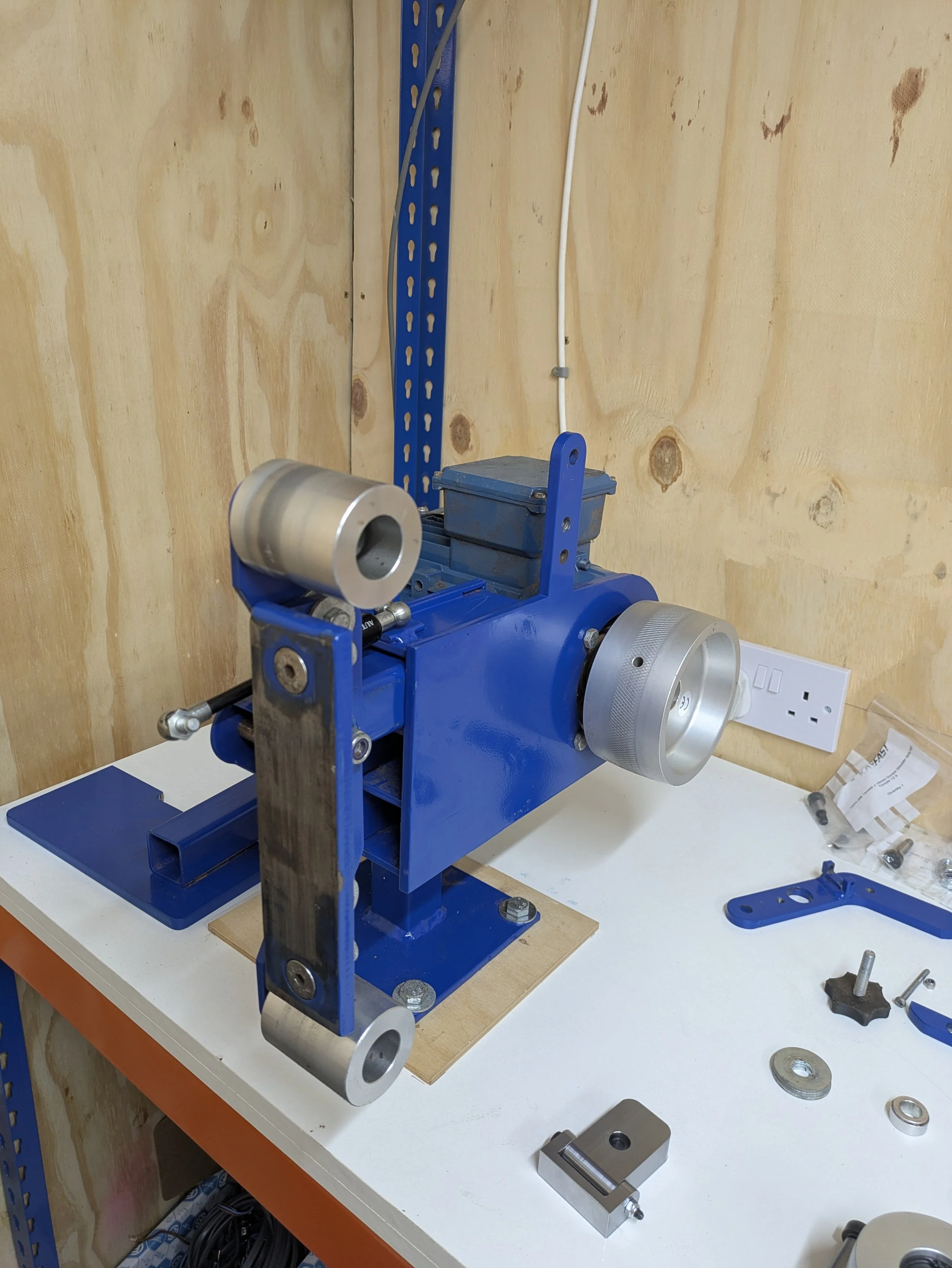 Tools and machinery on a workbench including a blue machine with silver rollers, a white electrical plug, and various small metal parts.