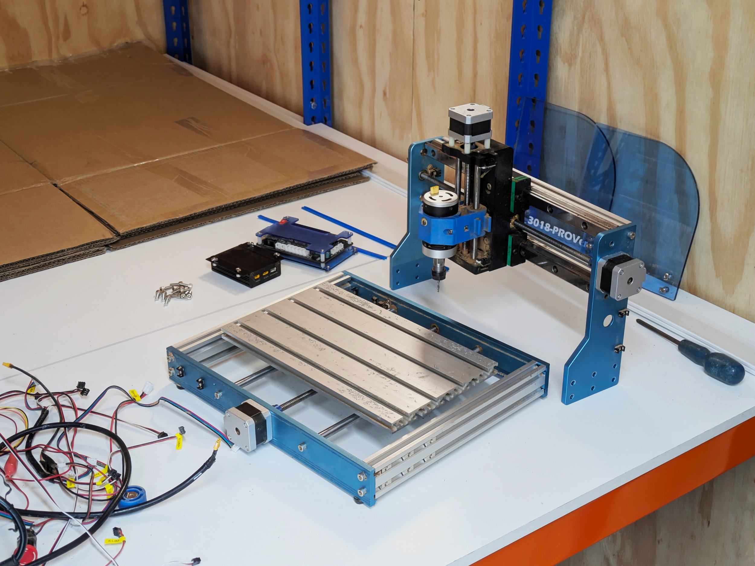A partially assembled CNC machine with various electronic components, wires, and cardboard pieces on a white work surface in a workshop.