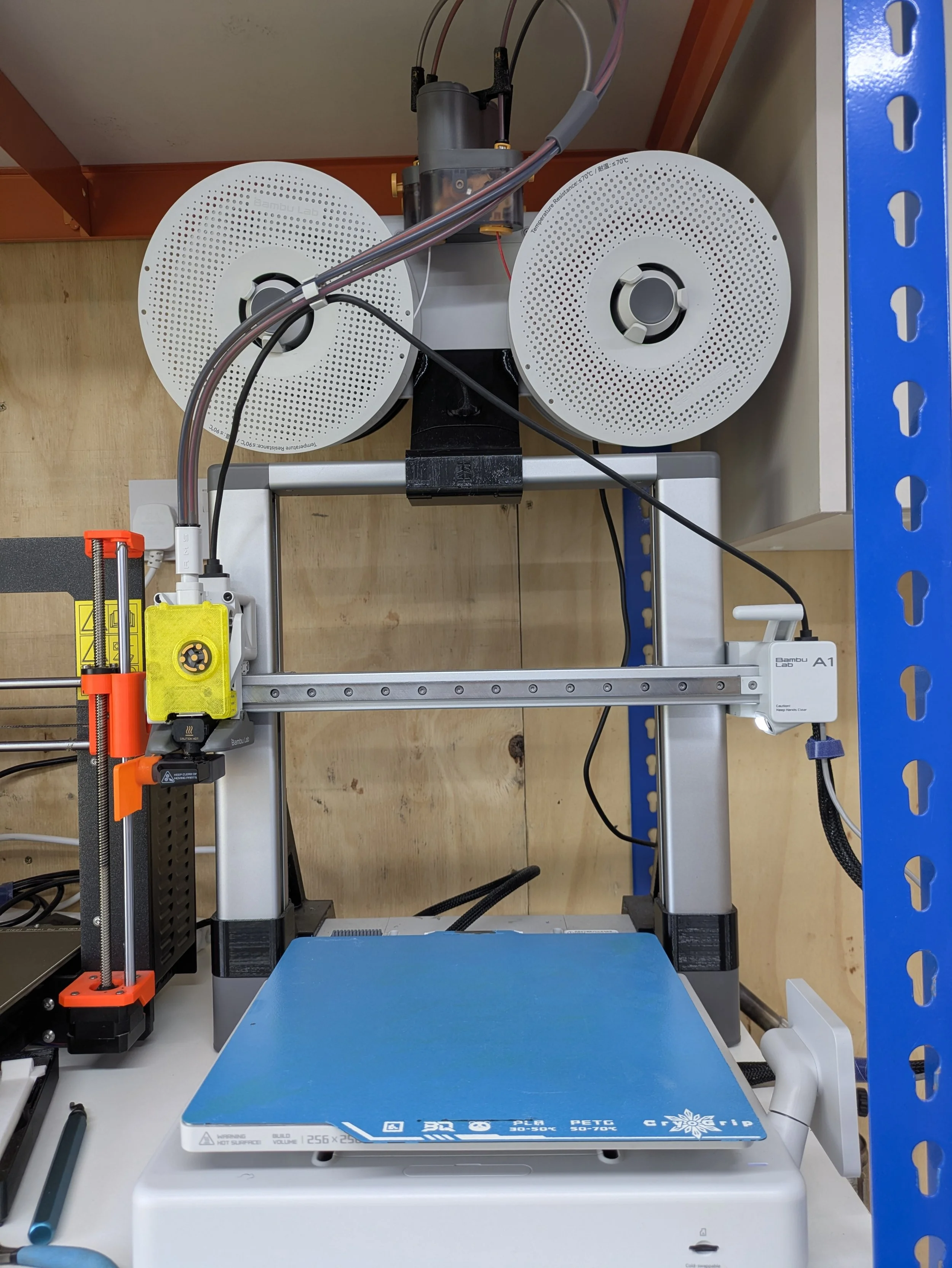 A 3D printer on a worktable in a laboratory setting, with a blue build surface, surrounded by wires and components.