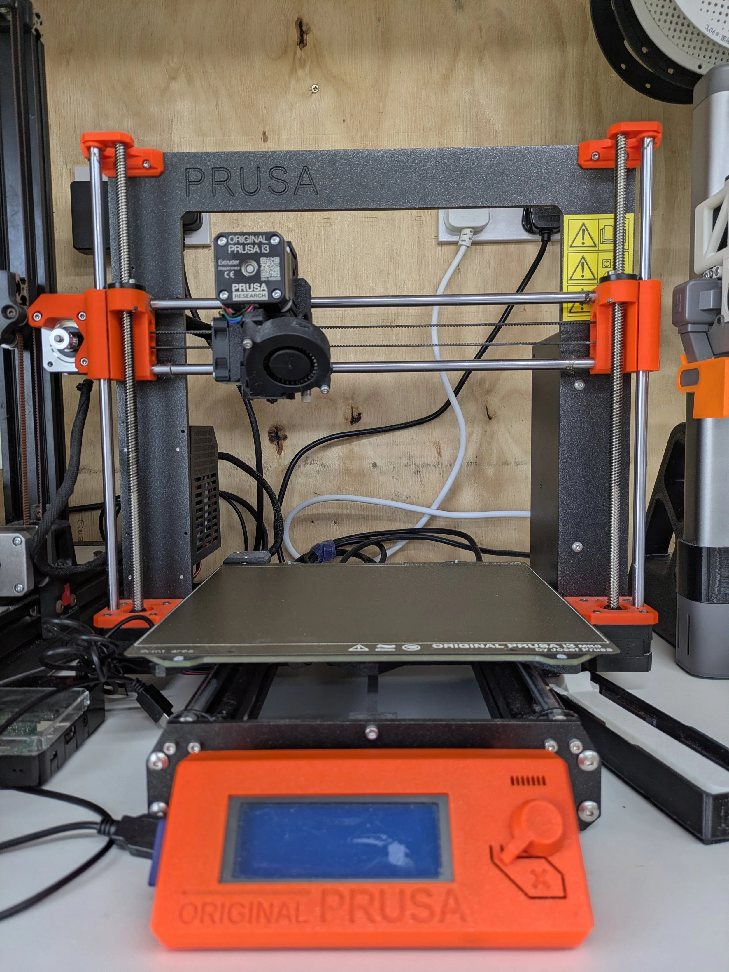 The image shows a 3D printer on a desk with its build platform in the foreground. The 3D printer has a black frame with orange components, and some wires and electronic parts are visible. The brand name "PRUSA" is engraved on the top frame of the printer.