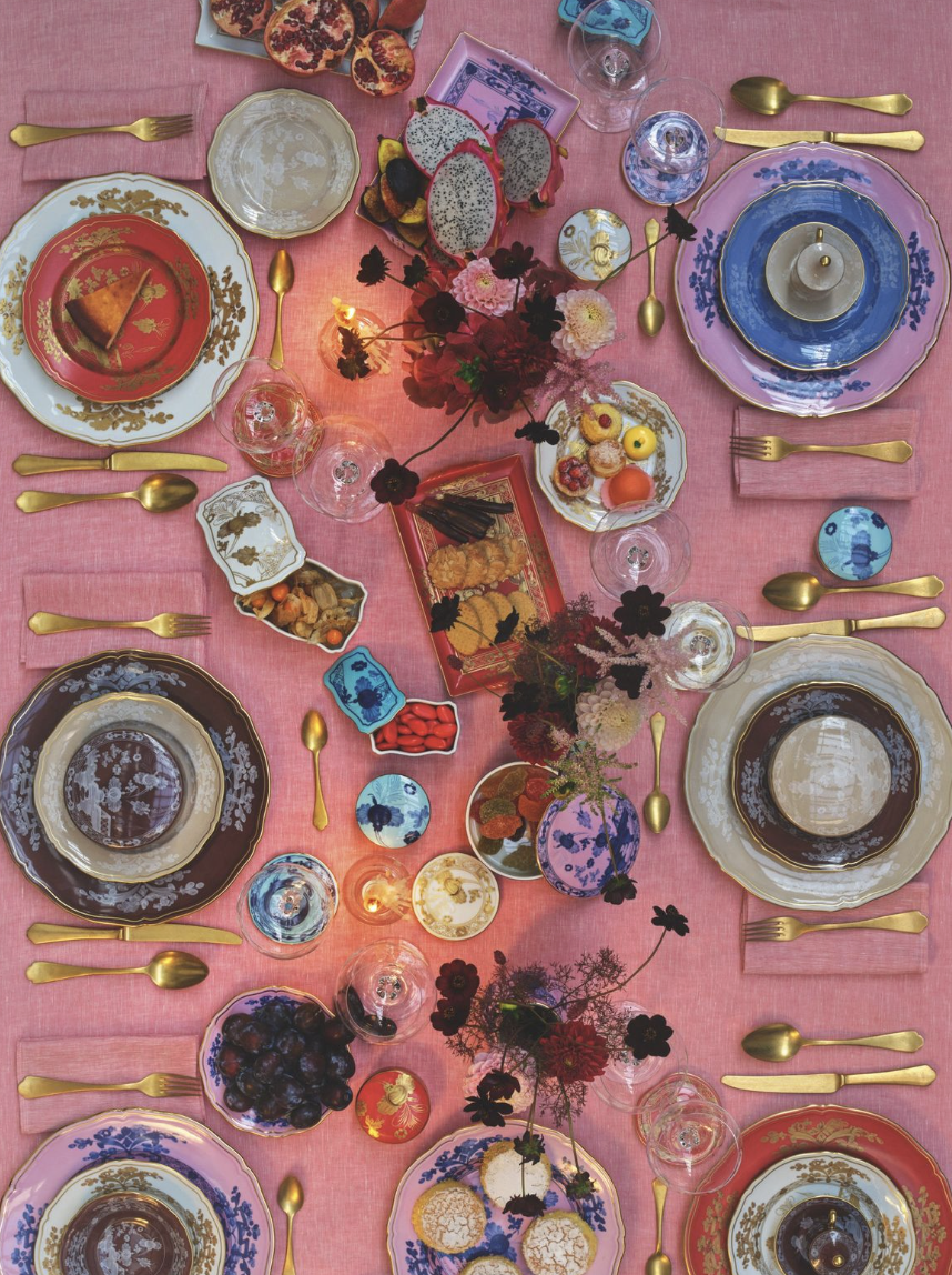 A festive table set with elegant china plates, gold utensils, and glasses, decorated with flowers, fruits, pastries, and candies on a pink tablecloth.