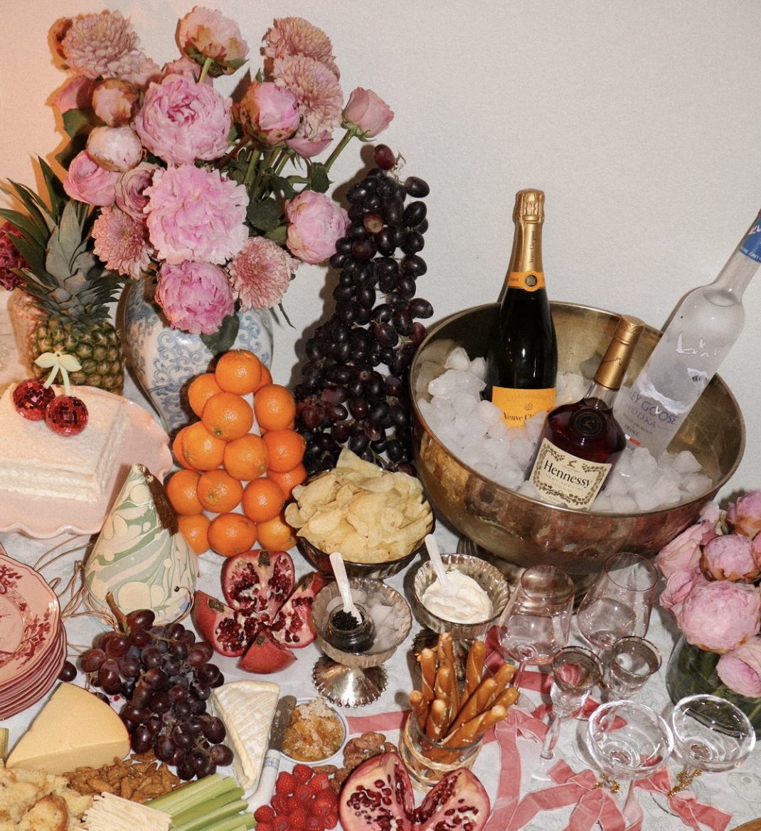 A table set with various fruits, cheeses, desserts, and drinks, including peonies and grapes in a floral vase, and a bowl of ice with bottles of champagne, vodka, and Hennessy.