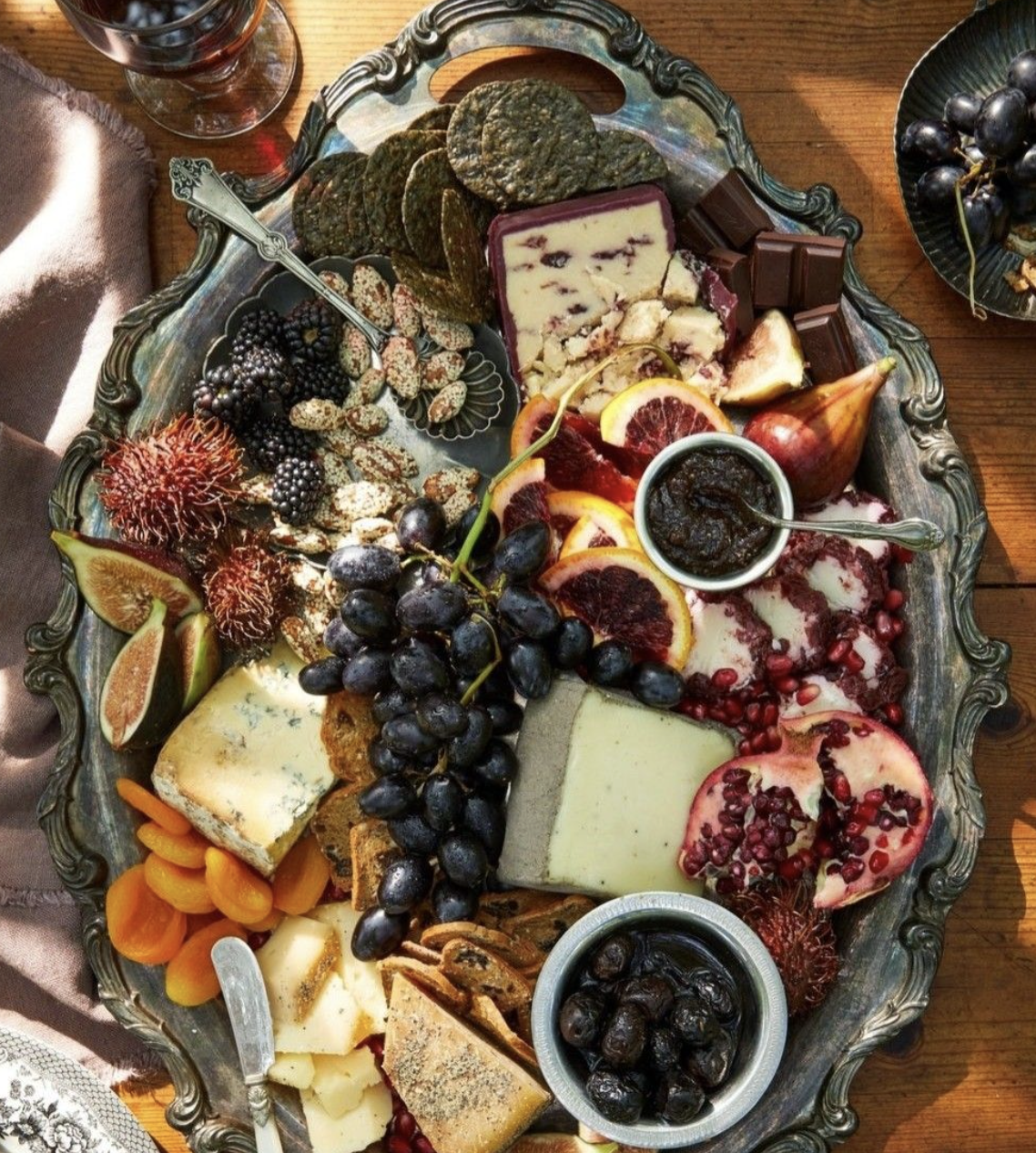 A large, ornate silver platter filled with assorted cheeses, grapes, figs, pomegranate seeds, chocolates, and preserved fruits.