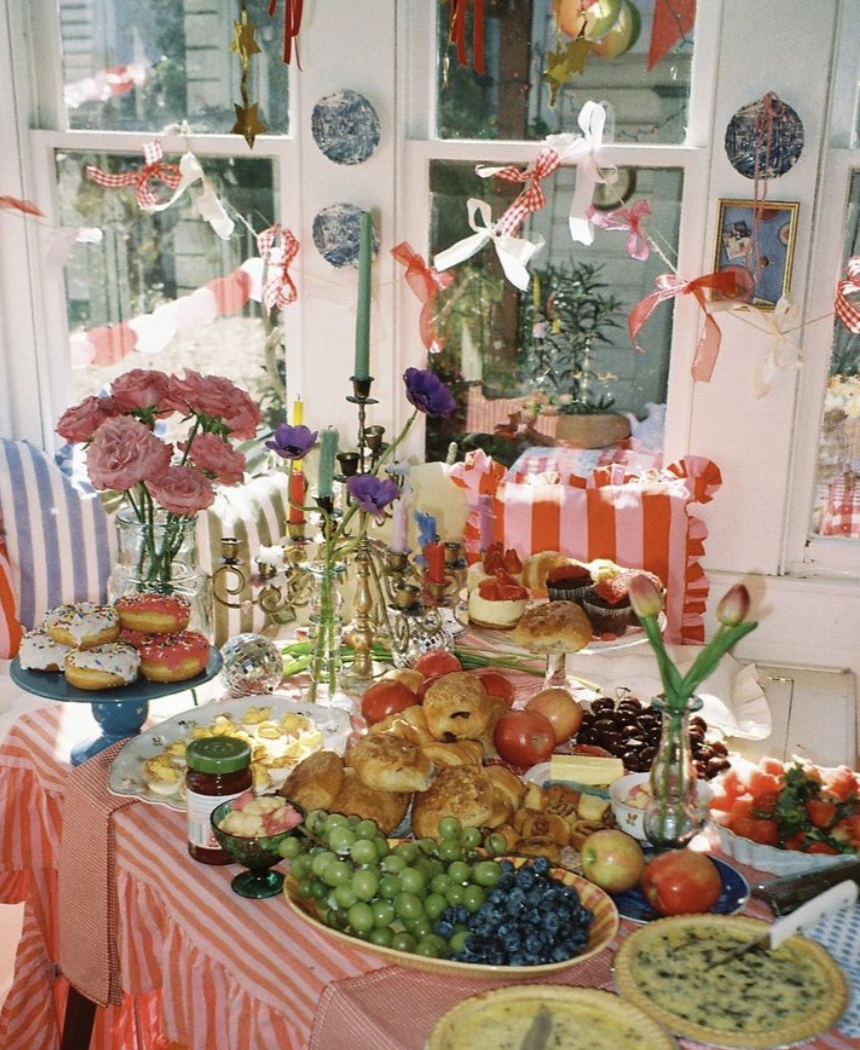 A table set for a celebration with various baked goods, fruits, flowers, and decorations, in front of a window decorated with ribbons and hanging ornaments.