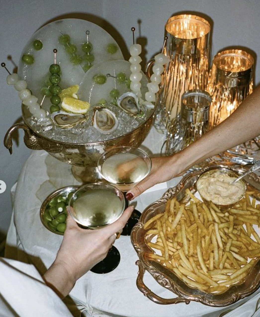 A woman and a man toasting with glasses of white wine at a social gathering. There is a large bowl of French fries with a side of dipping sauce, a bowl of green grapes, and a bowl of green and white olives on a table. The table is decorated with a bowl of ice containing oysters, lemon wedges, and skewers of green and white grapes. The background features a decorative rose gold lamp with several cylindrical shades.