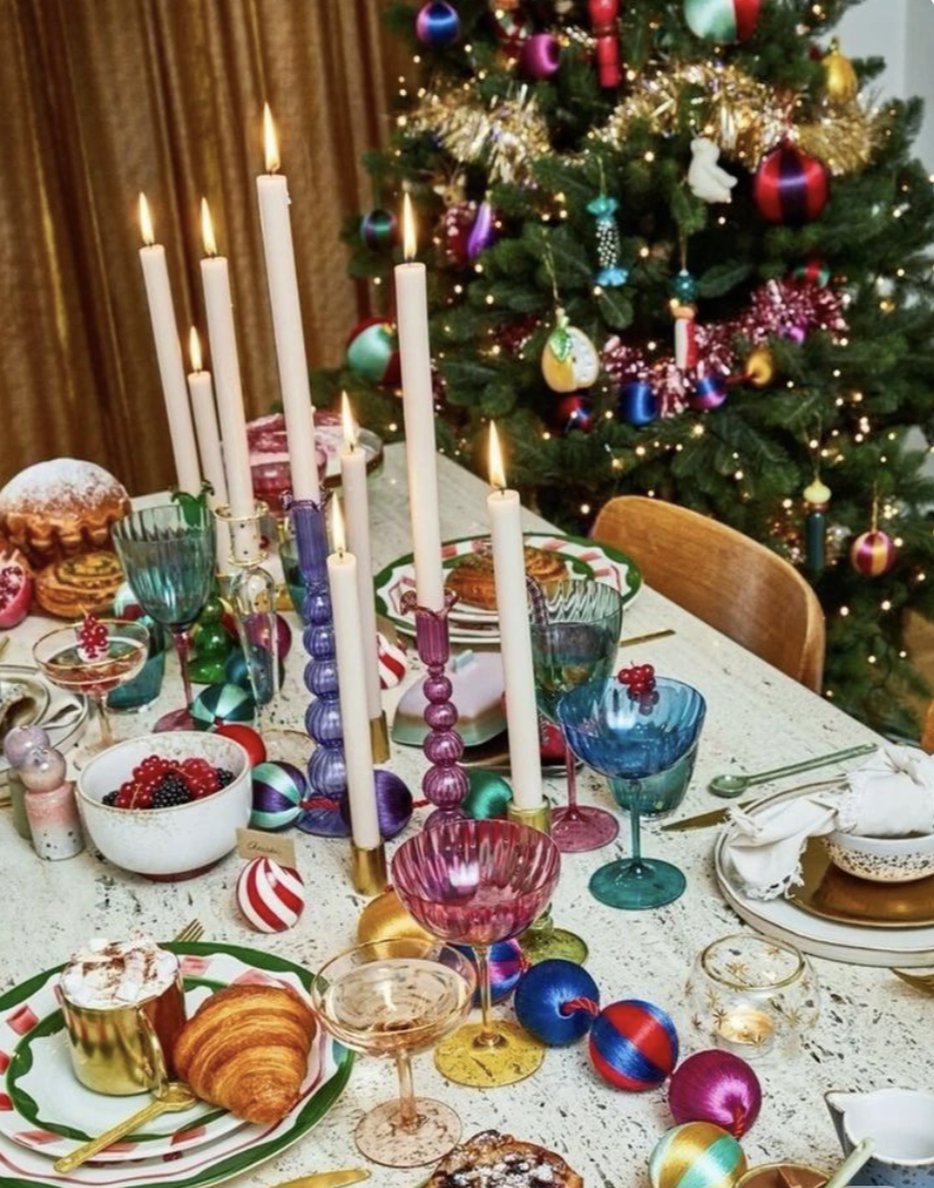 Festive Christmas table setting with colorful goblets, candles, and holiday treats, with a decorated Christmas tree in the background.