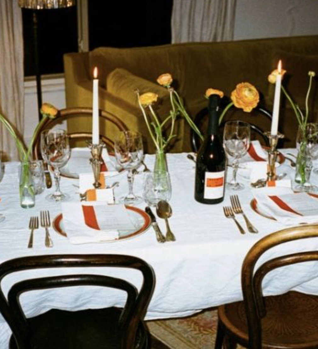 A dinner table set with six place settings, including plates, utensils, wine glasses, and napkins. There are four orange flowers in glass vases and two tall white candles. A bottle of red wine is placed at the center of the table. In the background, a green couch and window with curtains are visible.