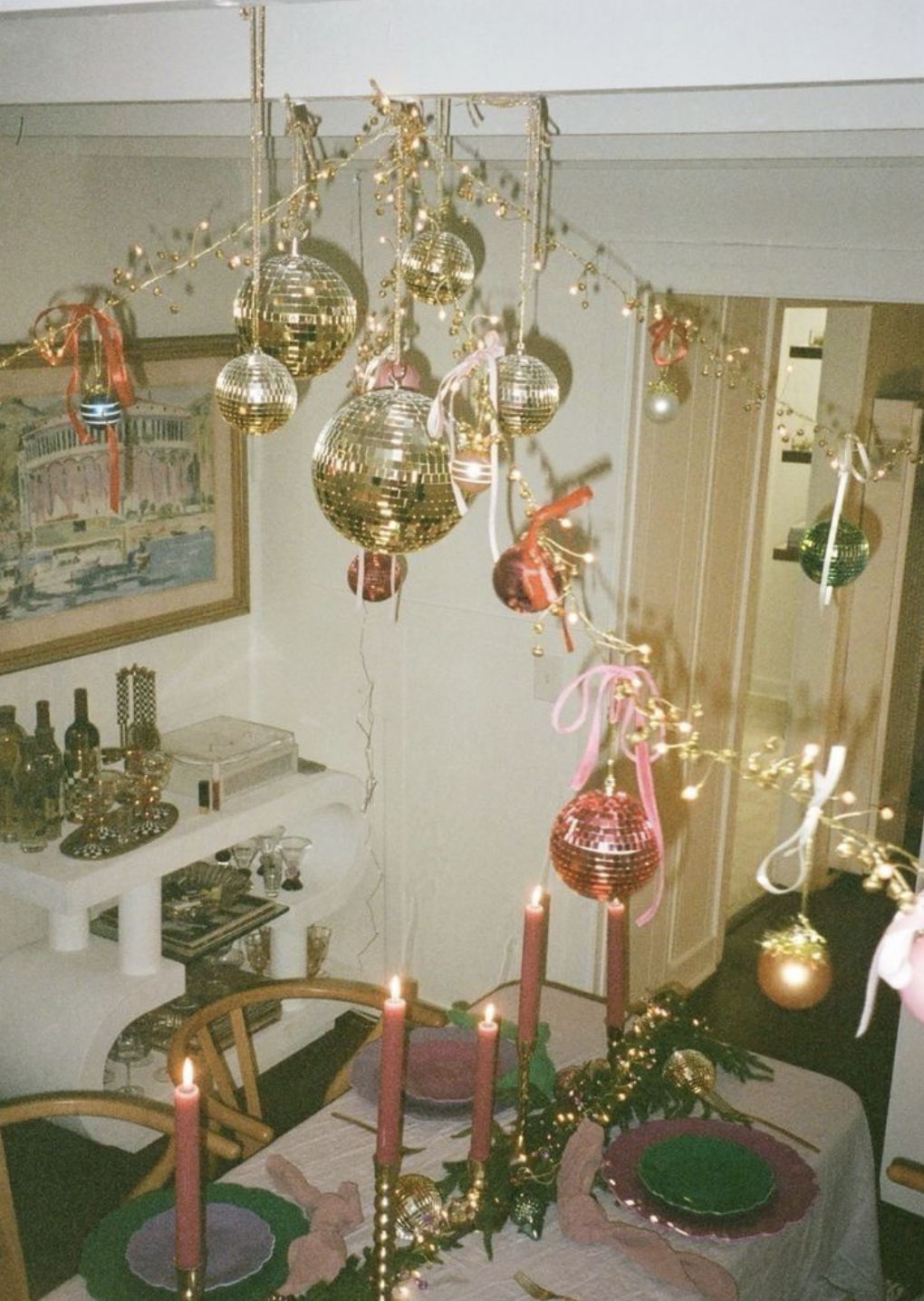 A dining room decorated for Christmas with hanging gold and pink ornaments, string lights, pink candles, and a Christmas garland on the table.