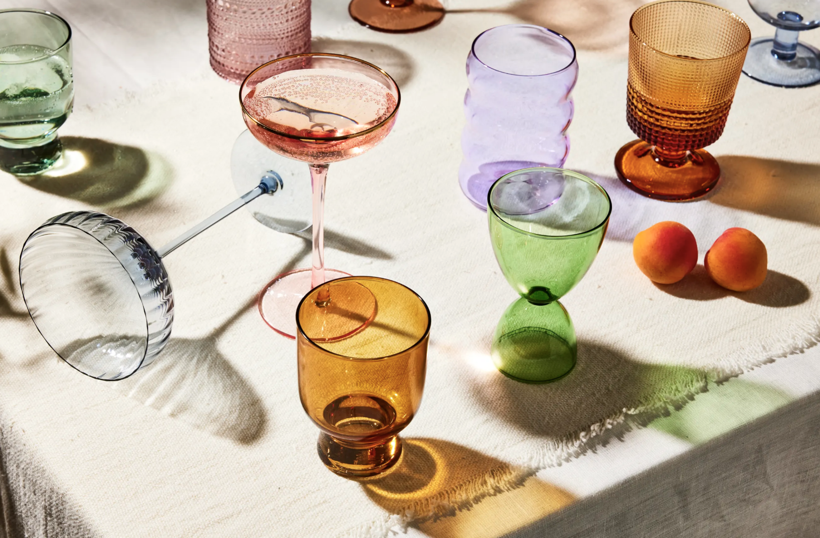 Colorful glassware including a pink cocktail glass, green, orange, purple, and blue drinking glasses, some with textured surfaces, along with two peaches on a white tablecloth.