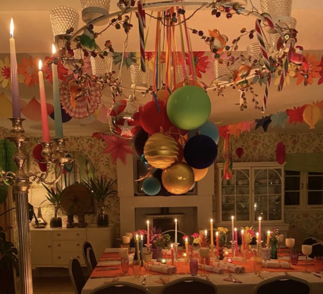 A decorated dining room with a large table set for a celebration, featuring lit candles, flower arrangements, and colorful decorations including balloons, paper ornaments, and hanging decorations on the ceiling.