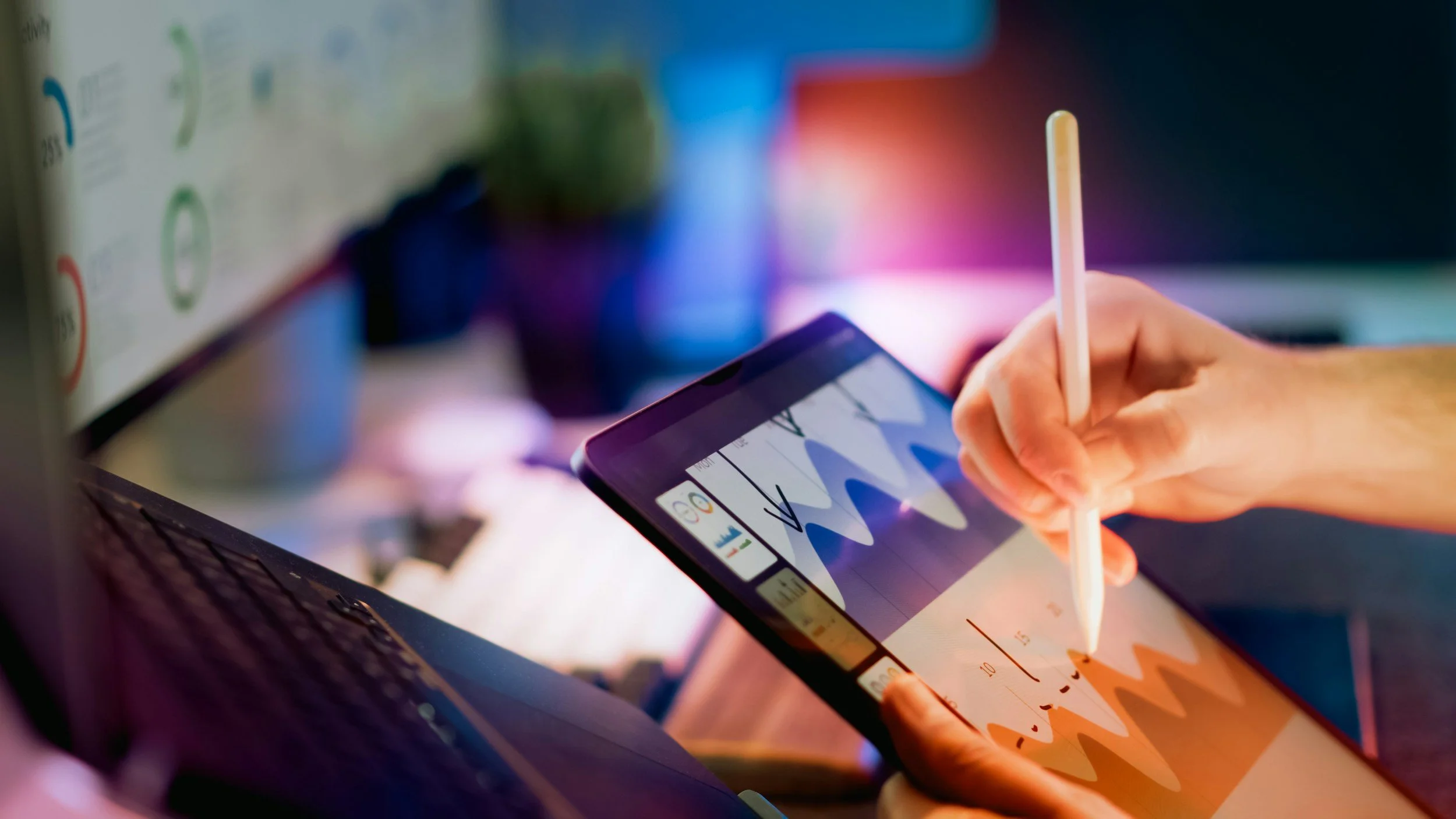 Person using a stylus on a tablet displaying colorful graphs and data charts, with a laptop in the background, under vibrant lighting.