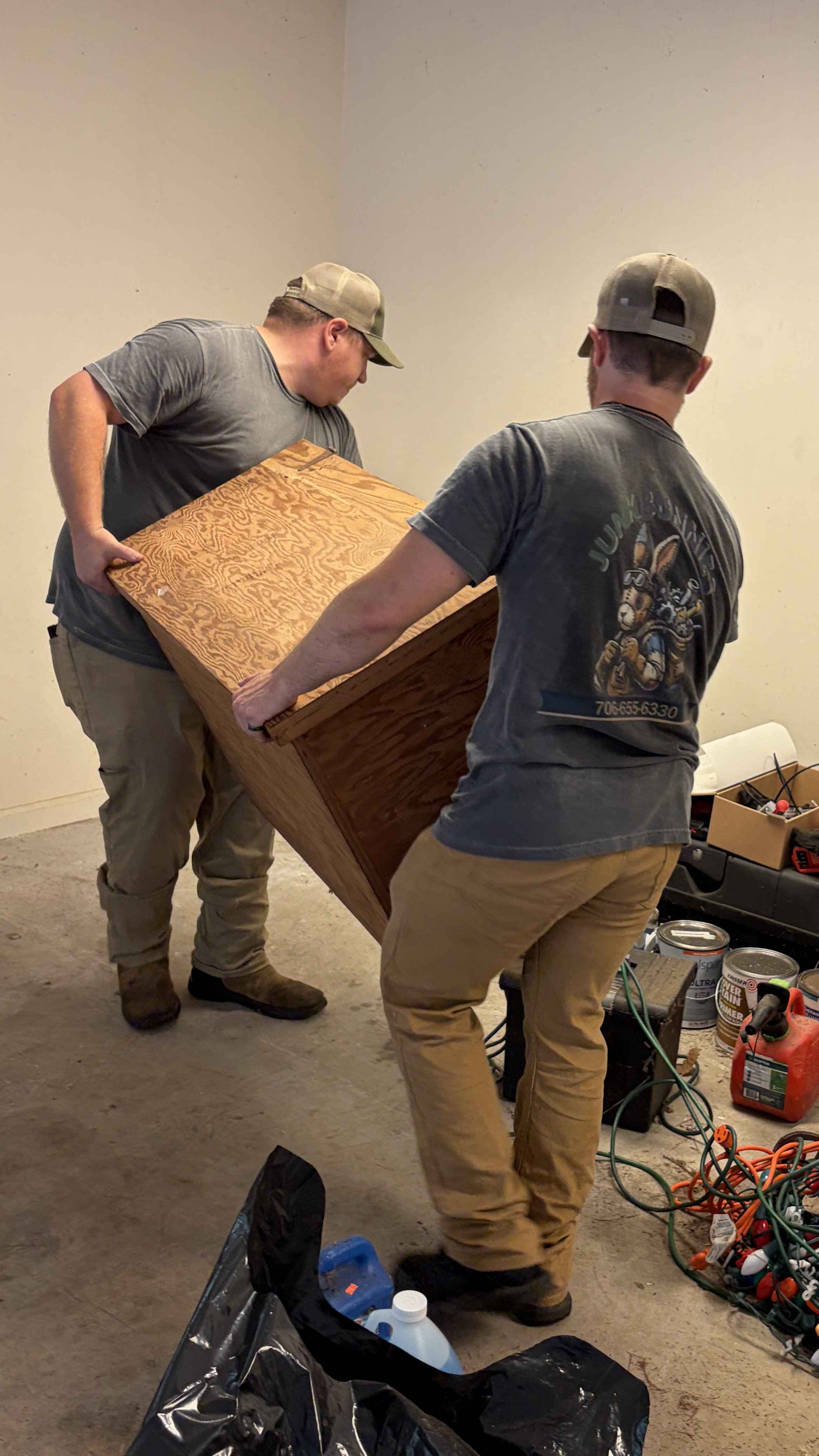 The Junk Bunnies are lifting a wooden cabinet in a room with a concrete floor and cluttered tools and supplies around.