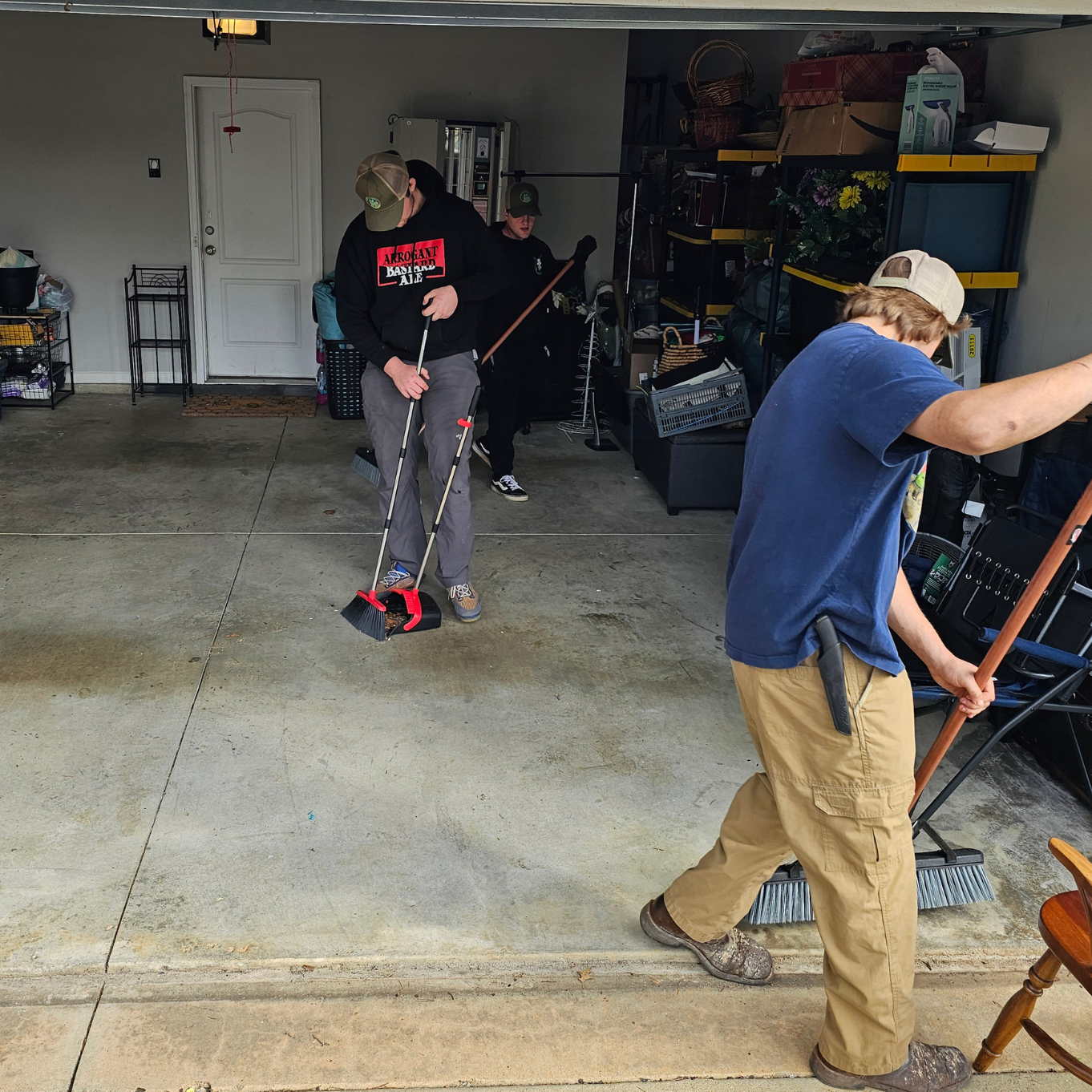 Three people cleaning a garage, sweeping and tidying up the space.