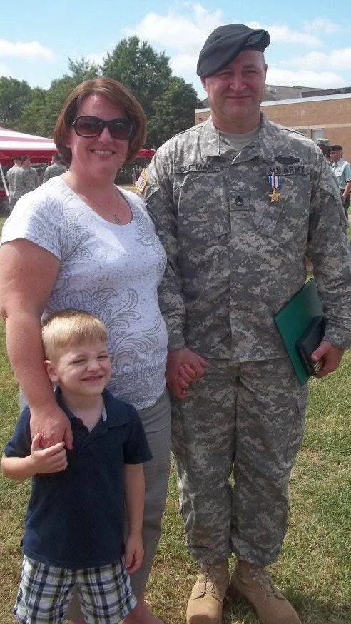 Staff Sergeant Sean Outman Stands in a field at an event with his Wife and Son. The woman and child are smiling. The Soldier Stands with a Silver star medal pinned on his Camouflage Uniform while he is wearing a black beret.
