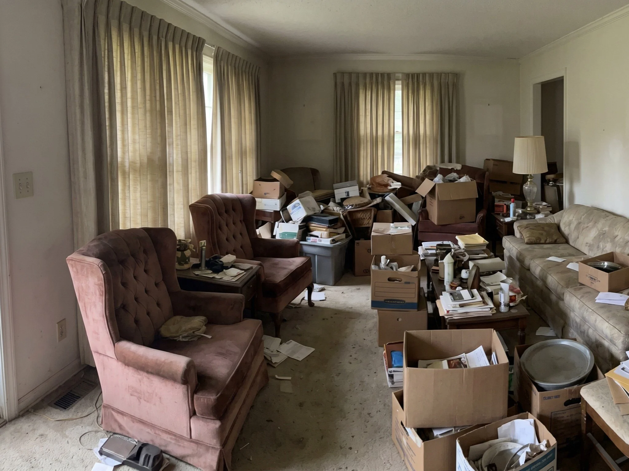 A cluttered living room with beige curtains, pink velvet armchairs, a floral sofa, floor and side tables, and a lamp. The room is filled with boxes, papers, and clutter.
