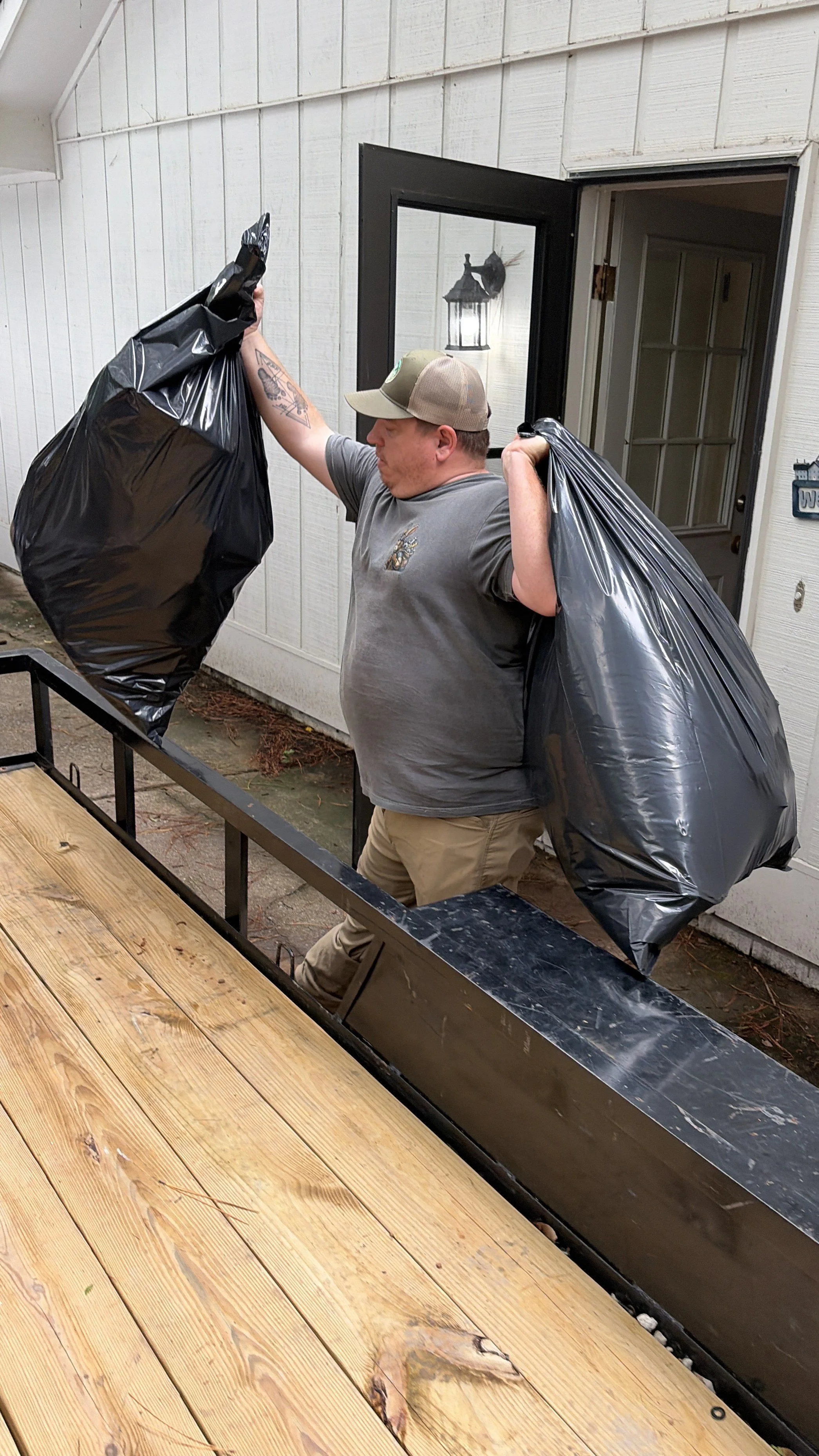 Man wearing a cap and gray shirt, carrying two large black trash bags, standing outside a house next to a wooden deck.
