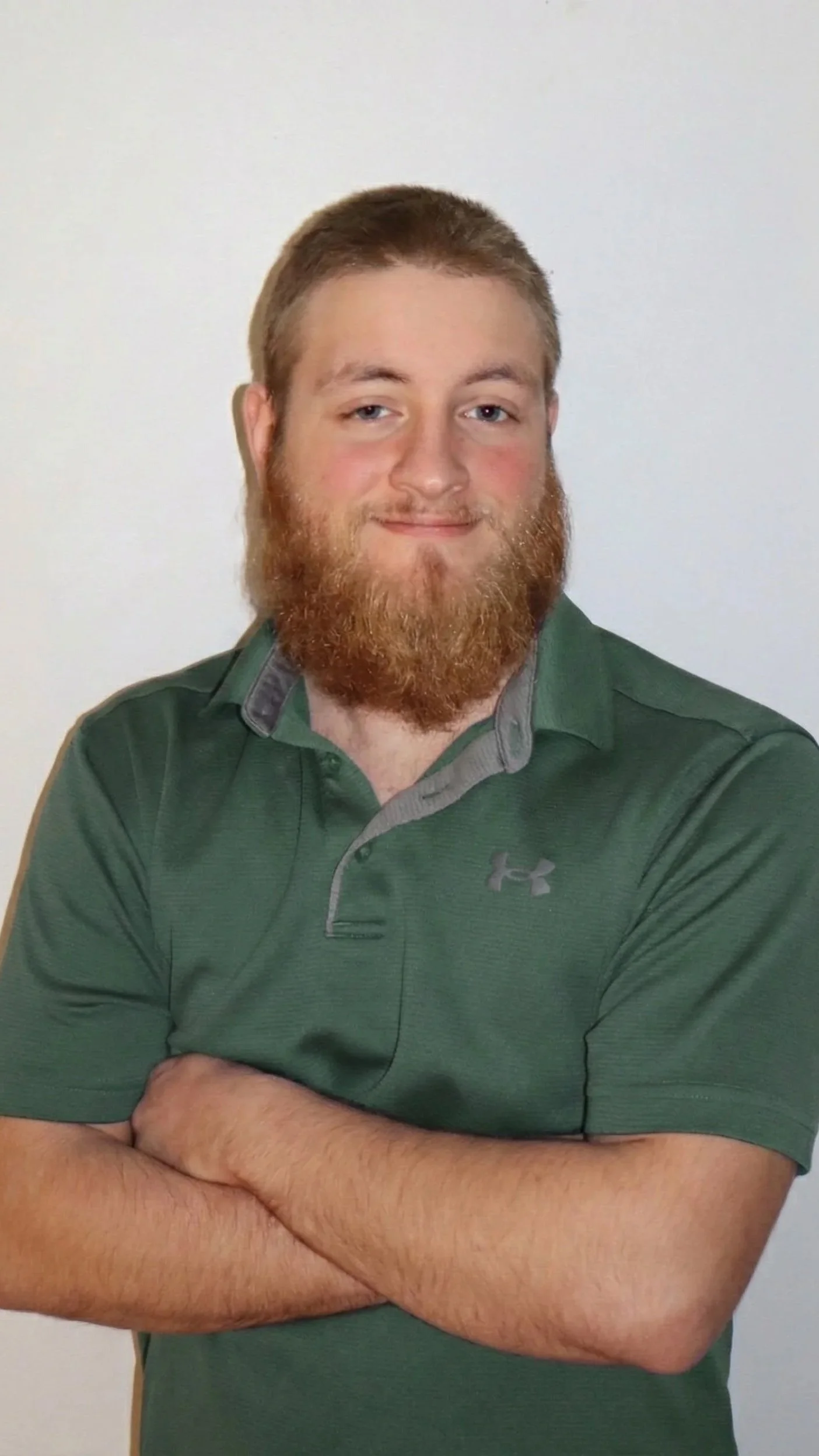 A young man with a beard and short hair, wearing a green polo shirt with a gray collar, standing with his arms crossed and smiling in front of a plain light-colored background.