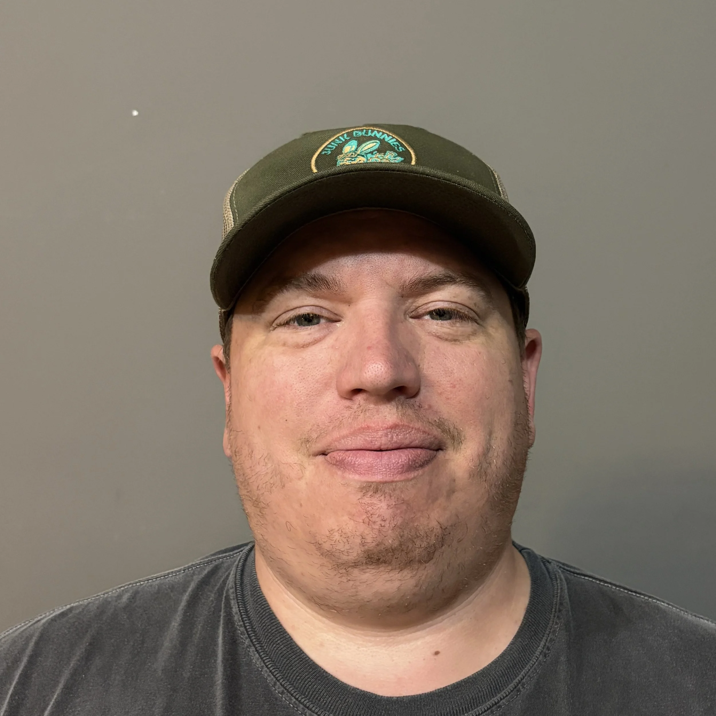 Close-up of Jacob Morgan Operations Manager of junk bunnies LLC wearing a green baseball cap with a Junk Bunnies Logo patch and a Grey shirt, against a plain gray background.