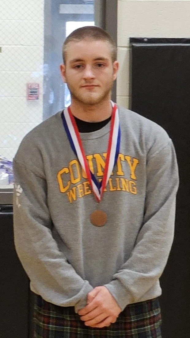 Michael Outman a Harris county Georgia wrestler with short hair wearing a gray sweatshirt with 'County Wrestling' printed on it, and a bronze medal around his neck, standing indoors in front of a black display or locker.