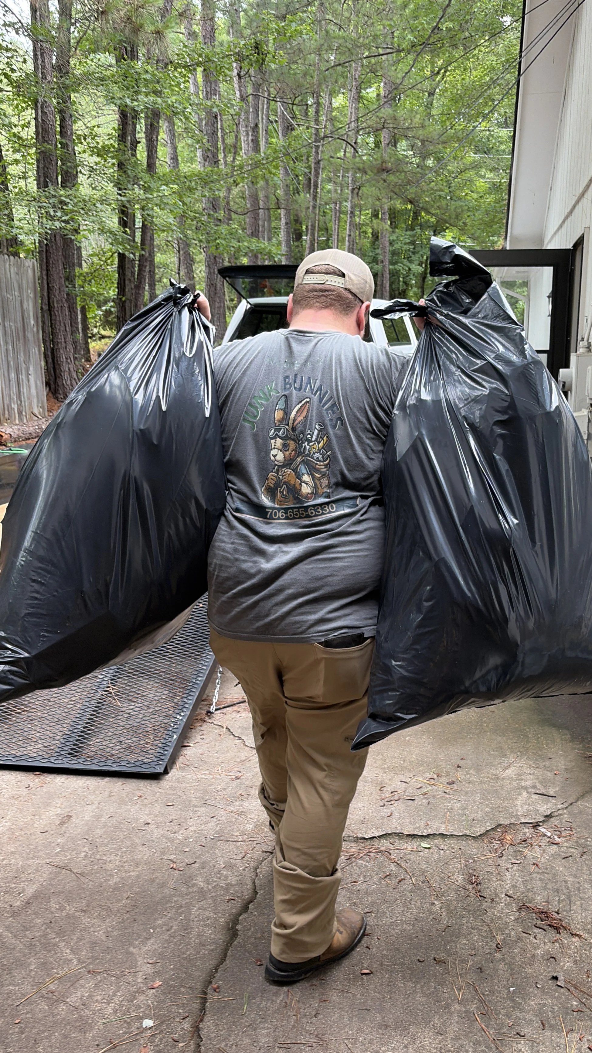 A man wearing a gray shirt with Junk bunnies graphic and khaki pants is carrying two large black trash bags outdoors near a wooded area. He is walking on a paved surface next to a building.