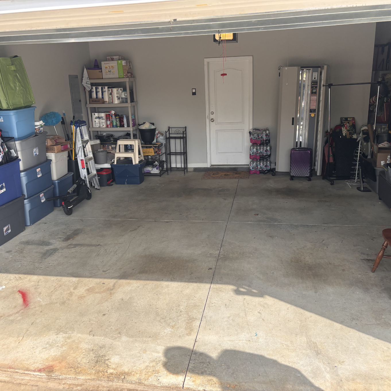 Cleaned and organized Garage interior with storage shelves, boxes, cleaning supplies, stacked bottled water, and a purple suitcase near an automatic door.