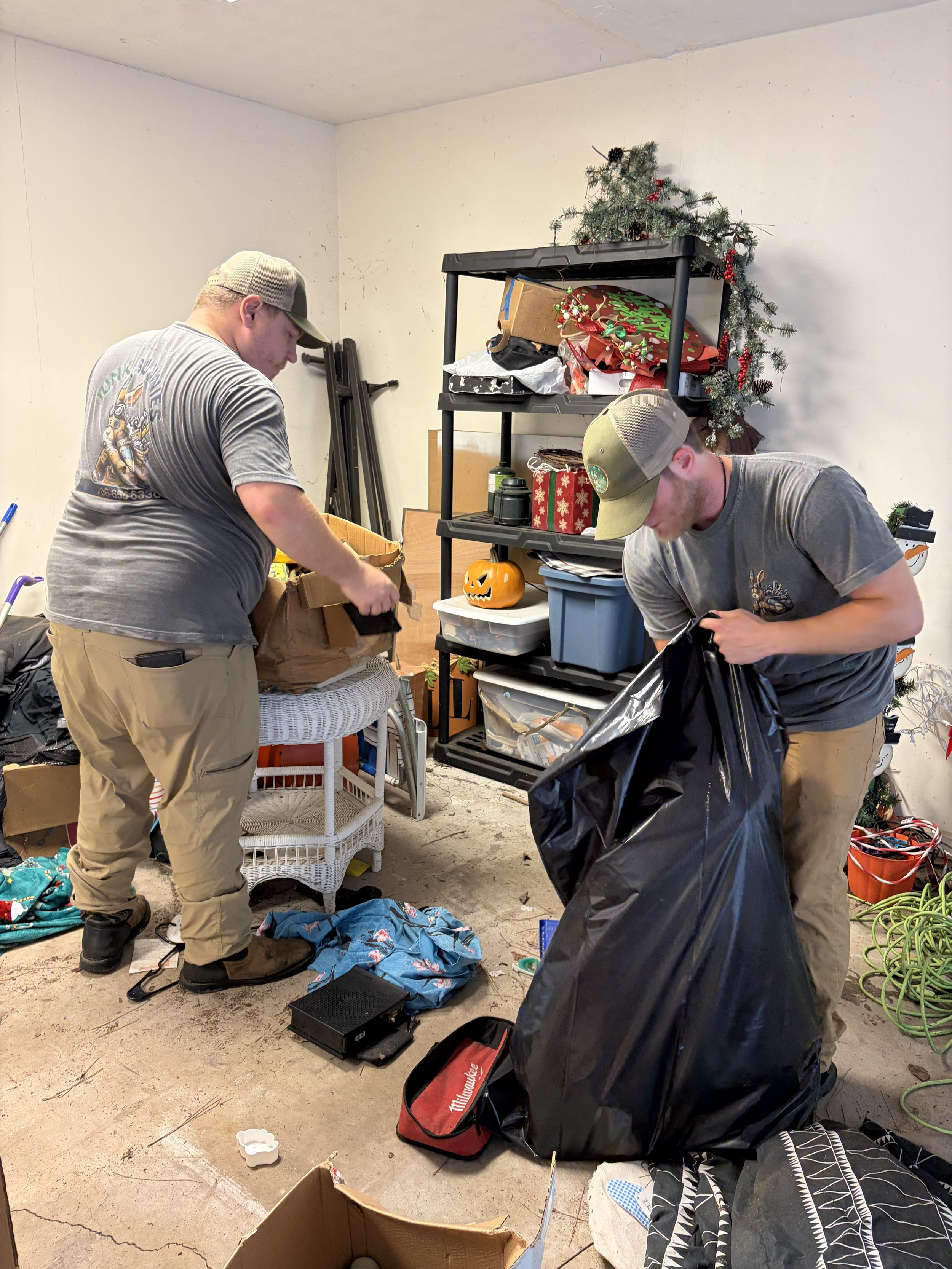 The Junk Bunnies organizing and packing items in a cluttered room decorated for Christmas, with a small Christmas tree on a shelf and a pumpkin decoration on a table.