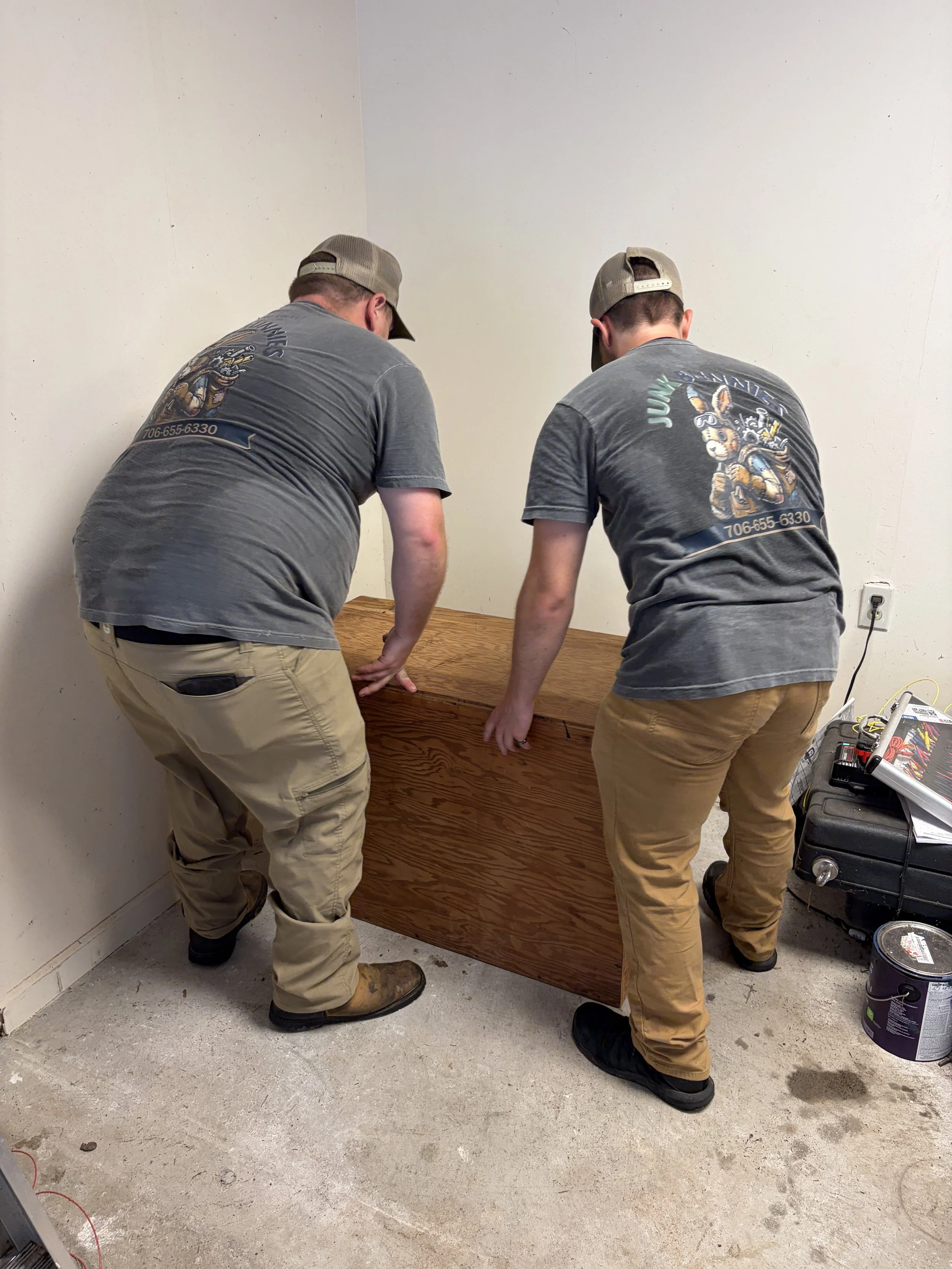 The Junk Bunnies are installing a wooden cabniet near a white wall and an electrical outlet, with tools and a bucket nearby.