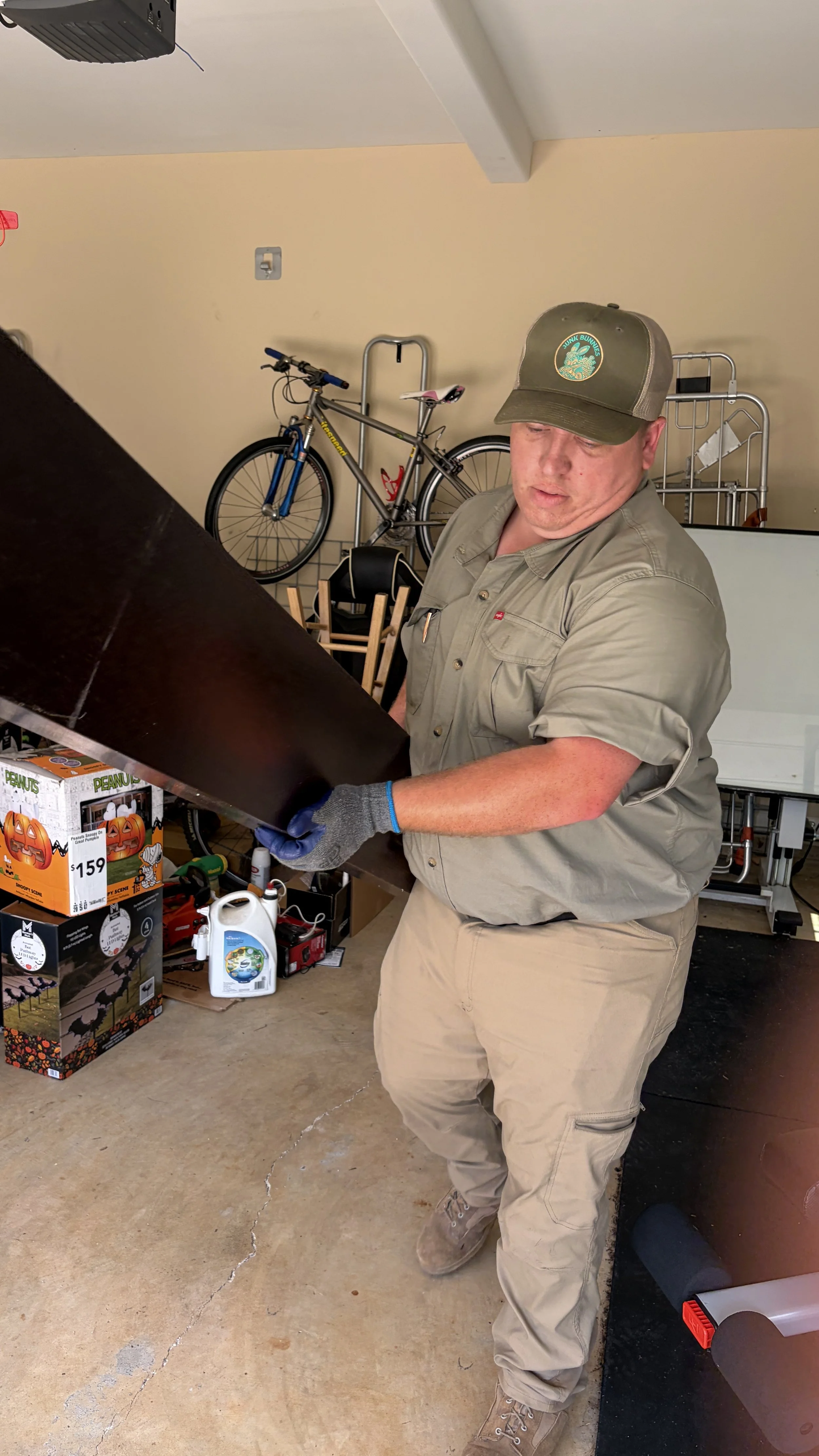 Jacob Morgan Junk Bunnies Operations manager is wearing a green baseball cap, khaki uniform, and gloves is working with a black panel in a garage. Behind him, there is a bicycle, a hand truck, boxes, and various objects stored in the garage.