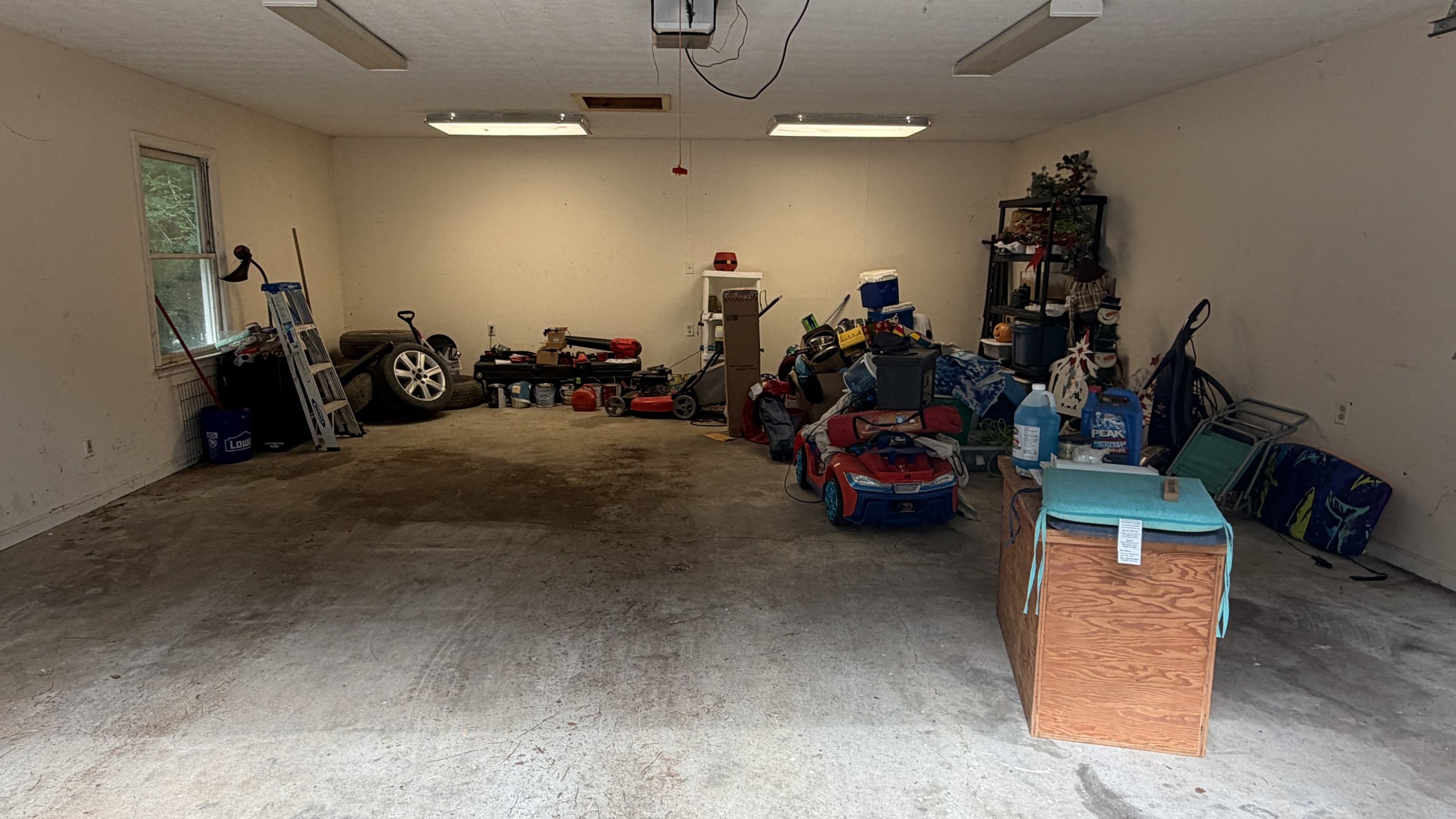 A cluttered garage with various items scattered, including a toy car, folding chairs, storage bins, a workbench, and miscellaneous household items.