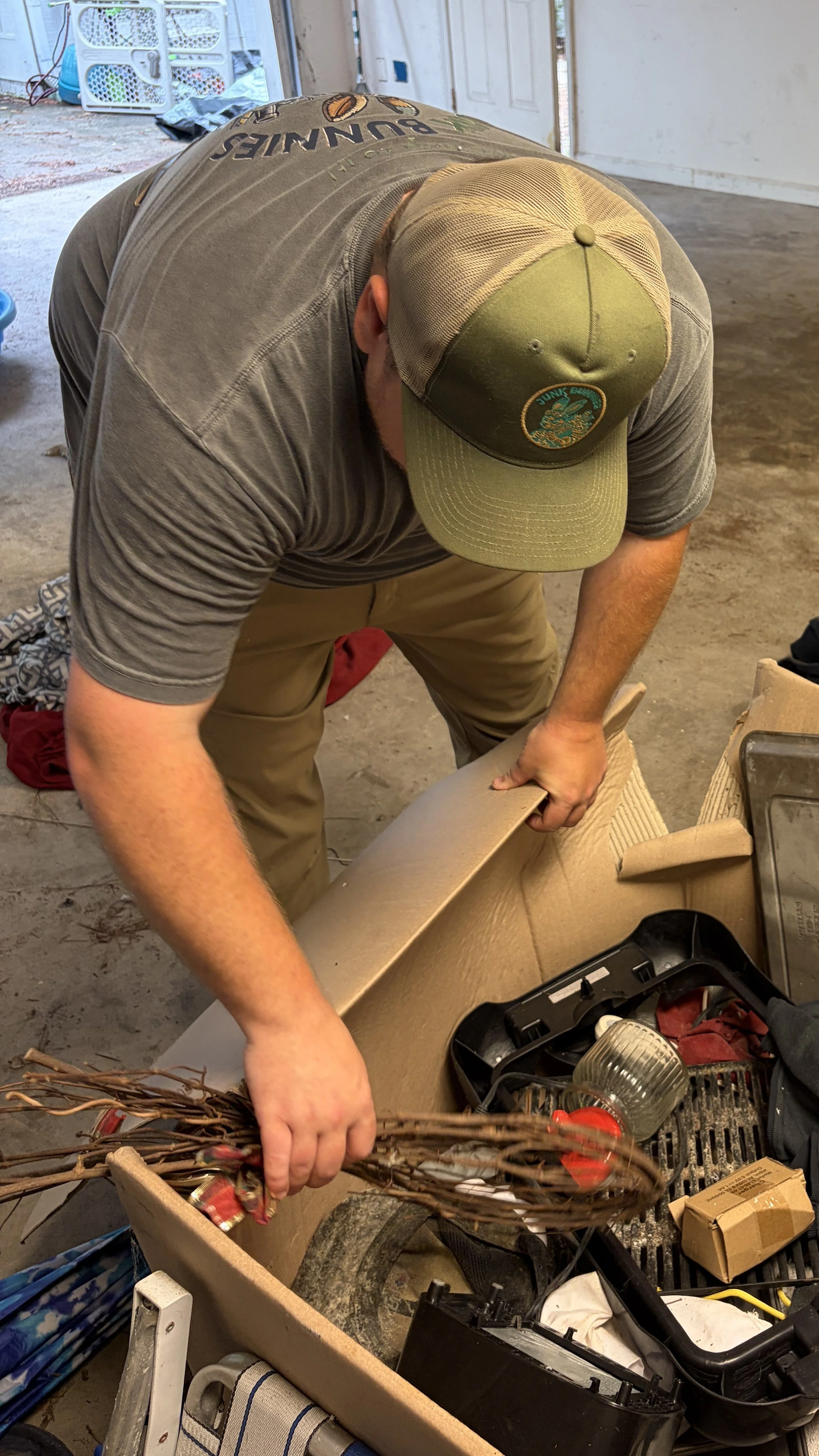A person wearing a green cap with a Junk Bunnies logo, gray t-shirt, and tan pants is working on assembling or fixing a cardboard structure inside a garage or workshop. There are various tools, boxes, and miscellaneous items around.