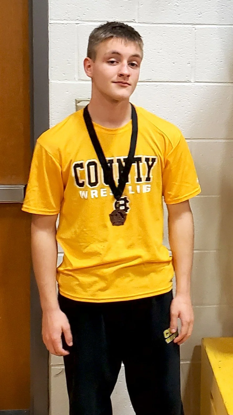 Young male wrestler wearing a yellow T-shirt with 'Harris County Wrestling' printed on it, a medal around his neck, and black sweatpants standing indoors against a cinder block wall.