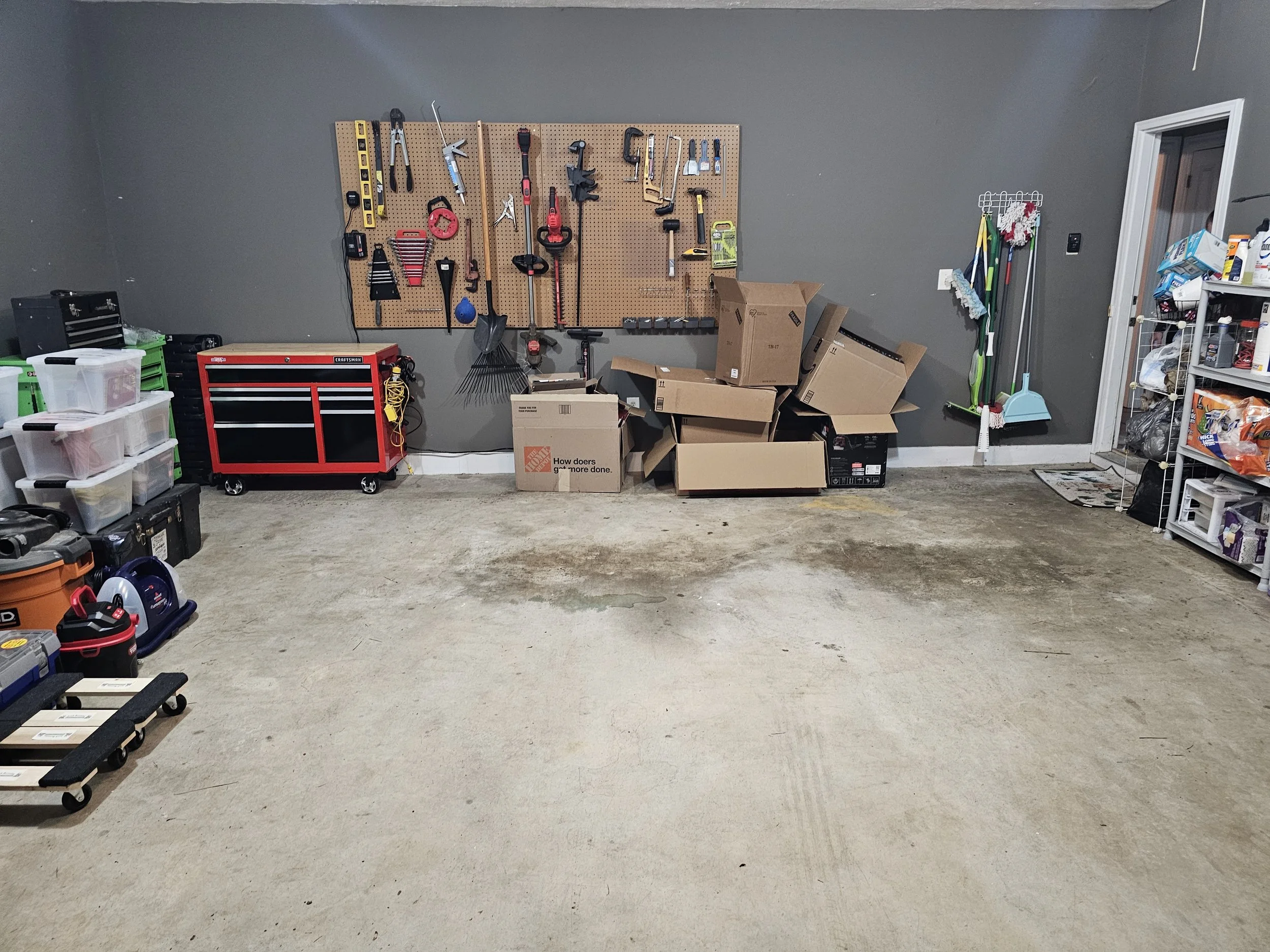 A neatly organized garage with a pegboard wall holding various tools and supplies, a red tool chest, plastic storage bins, cardboard boxes, and cleaning tools.