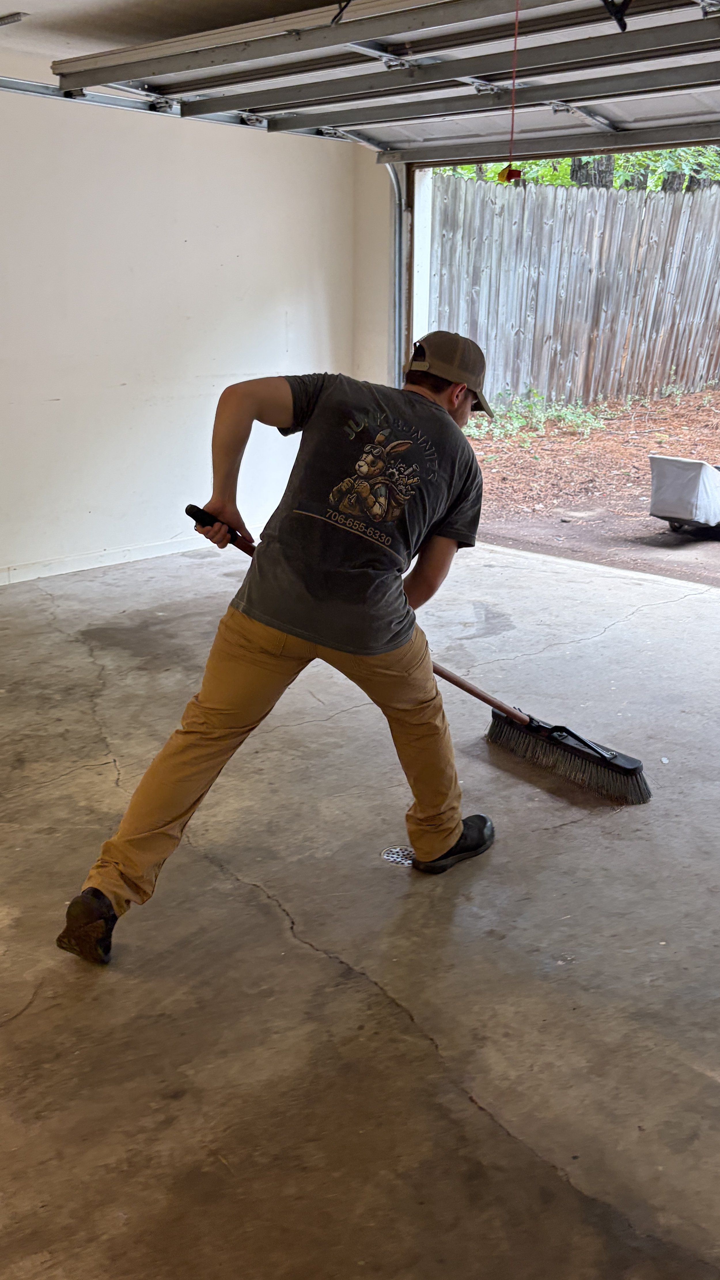 Michael outman The owner of Junk Bunnies sweeping concrete floor inside a garage with a broom, wearing tan pants, a dark gray T-shirt, and a baseball cap.