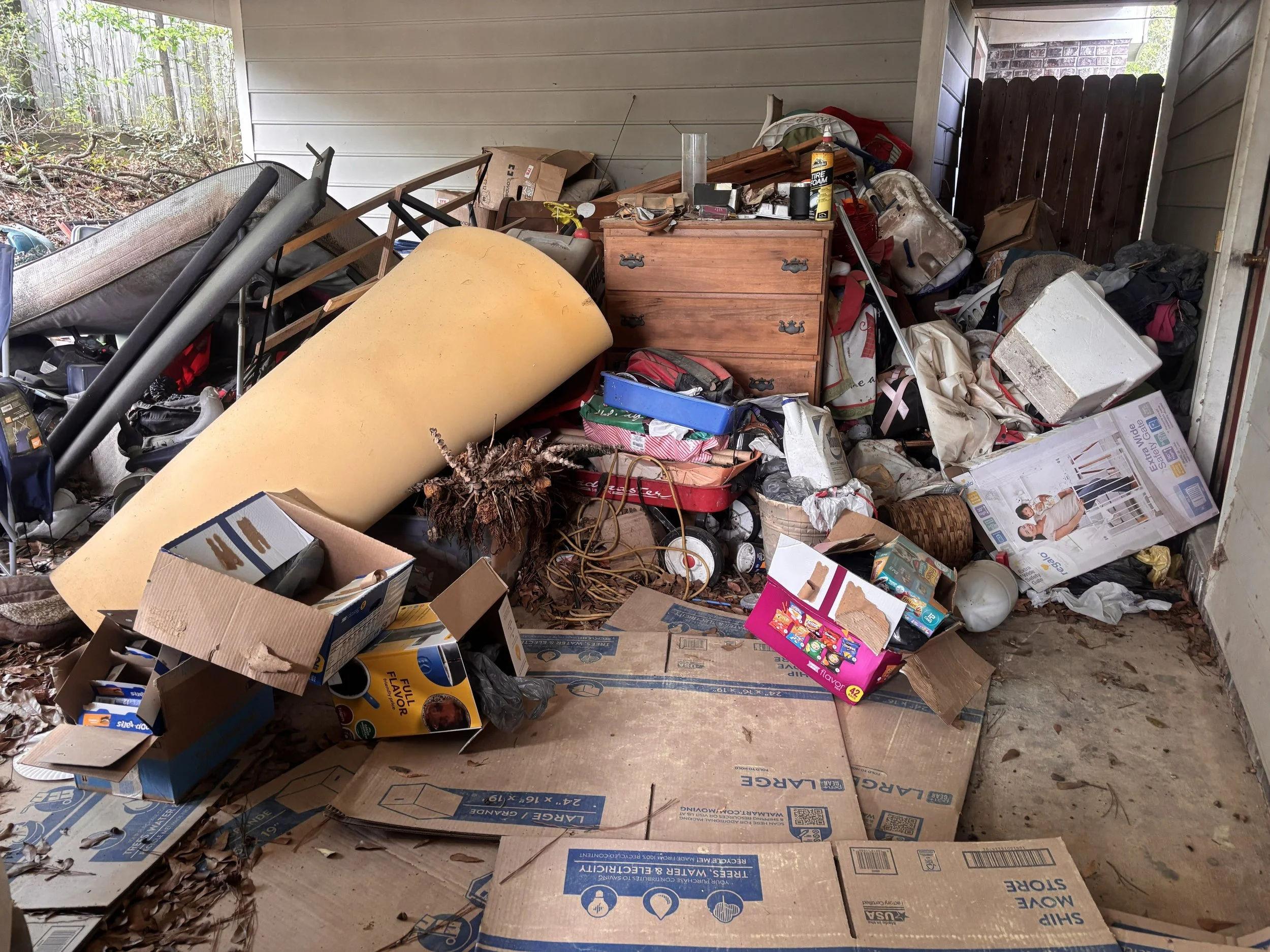 A cluttered outdoor porch or garage filled with boxes, furniture, and miscellaneous household items.
