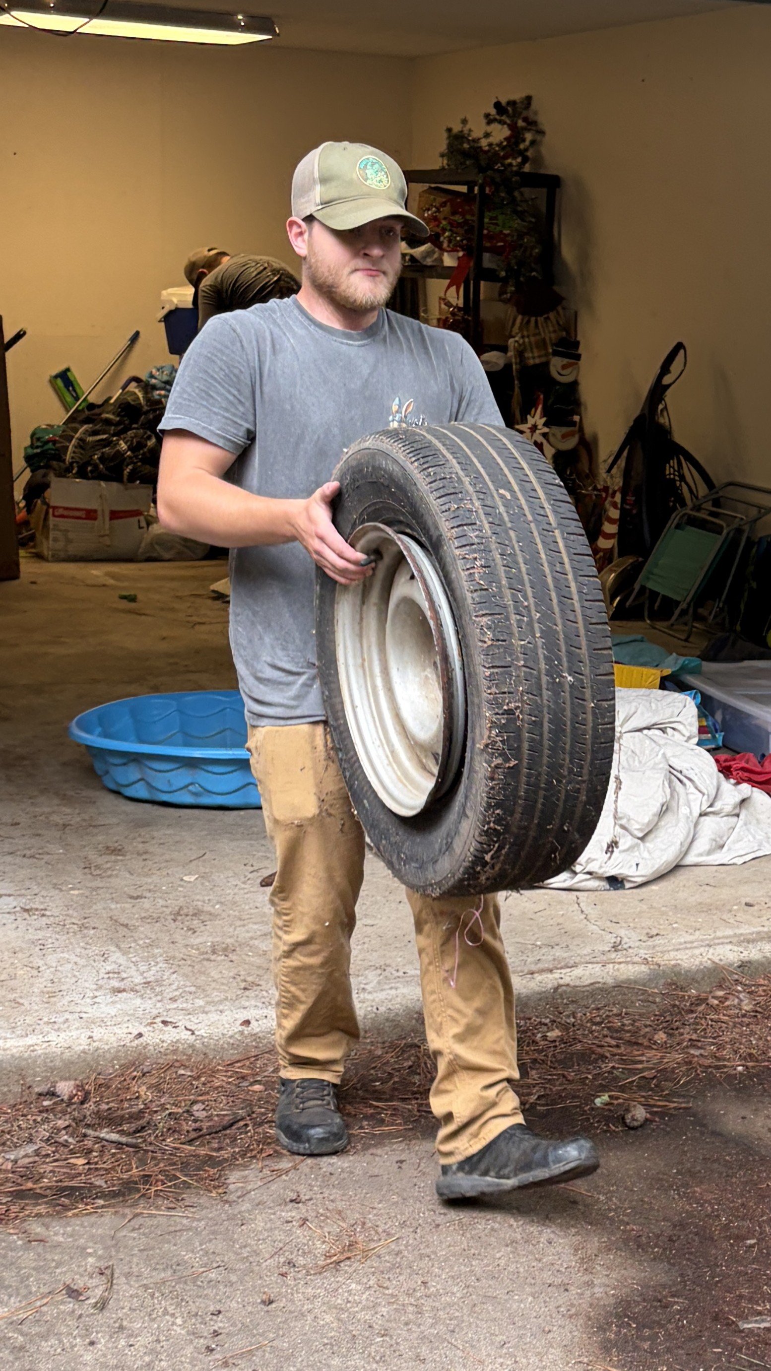 Michael outman The owner of Junk Bunnies wearing a gray T-shirt, tan pants, and black boots holds a large, worn tire indoors with a cluttered background, including a blue plastic tub, a bag of items, and some shelves.