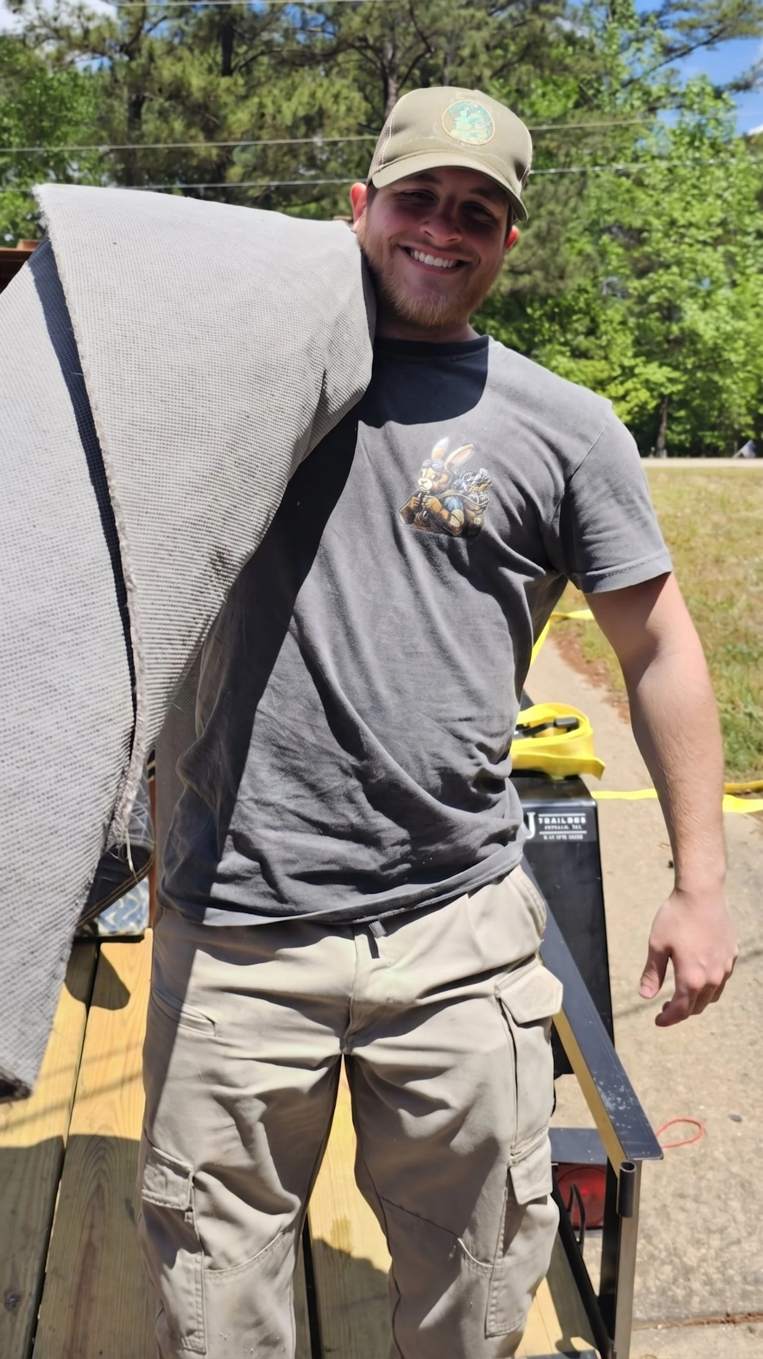A man smiling outdoors, wearing a gray t-shirt with a cartoon rabbit on it, beige cargo shorts, and a beige hat with a logo, standing on a wooden deck with trees in the background.